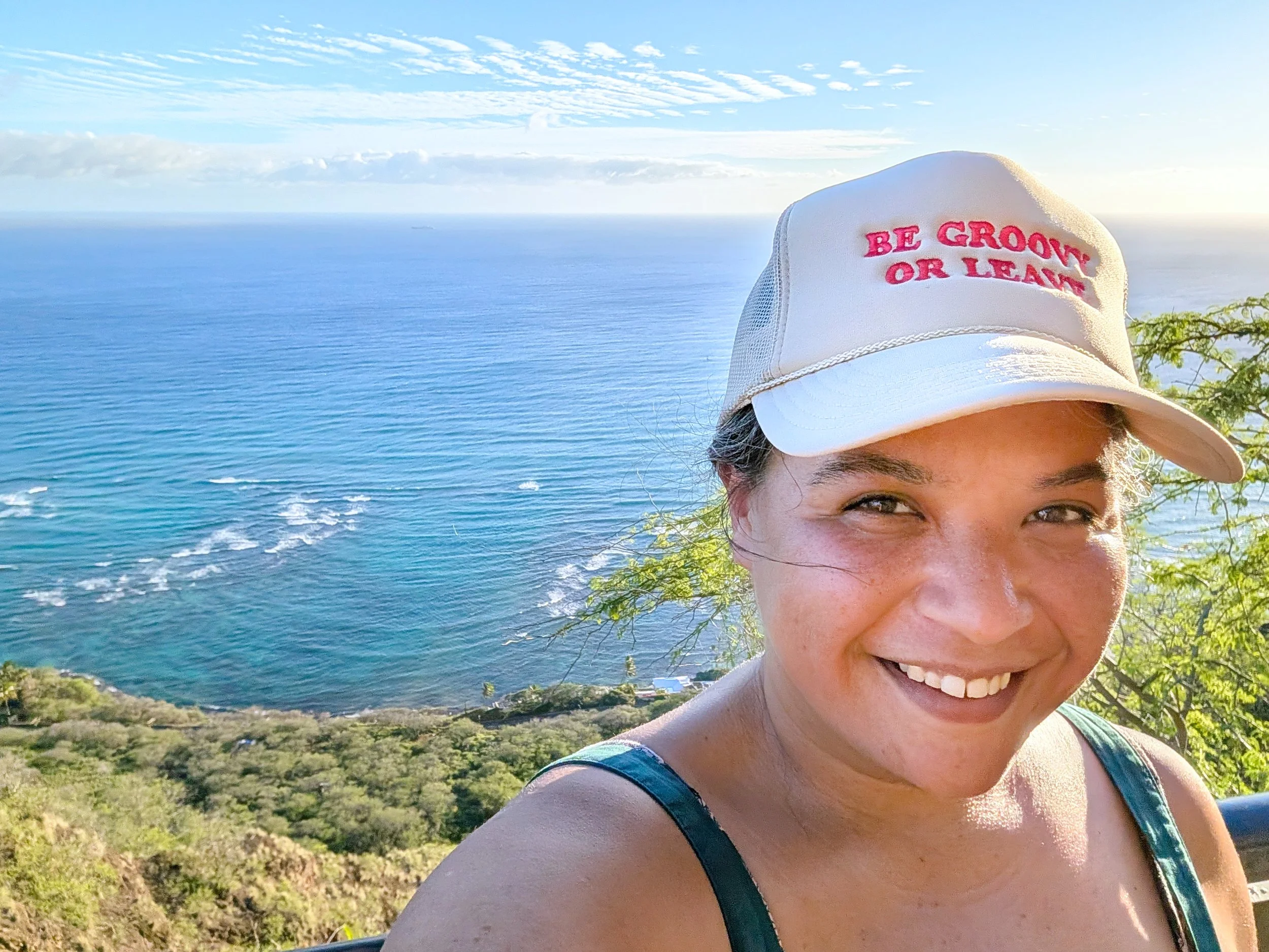A woman smiling outdoors near the ocean, wearing a white baseball cap with pink embroidered text that reads 'BE GROWN OR LEARN'. The background shows blue ocean water, a cloudy sky, and greenery on the hillside.