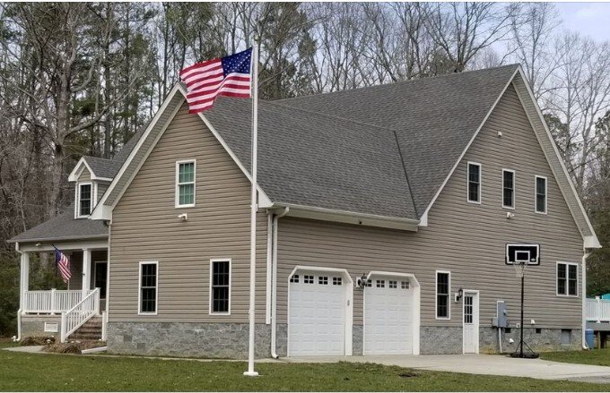 Two-story beige house with a gray roof, American flag on a pole, basketball hoop, and a multi-car garage, surrounded by trees.
