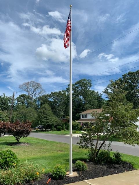 American flag on a tall flagpole in a residential yard with trees and a house in the background under a blue sky with clouds.