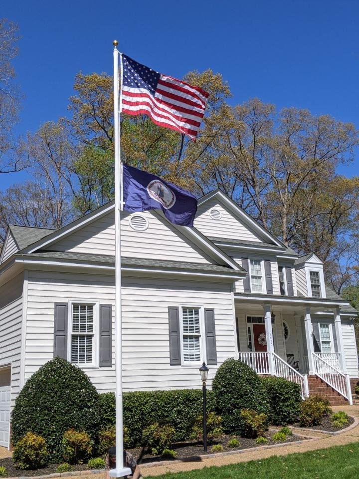 House with American and Virginia flags on flagpole, white facade, front porch, neatly trimmed hedges, and clear blue sky.