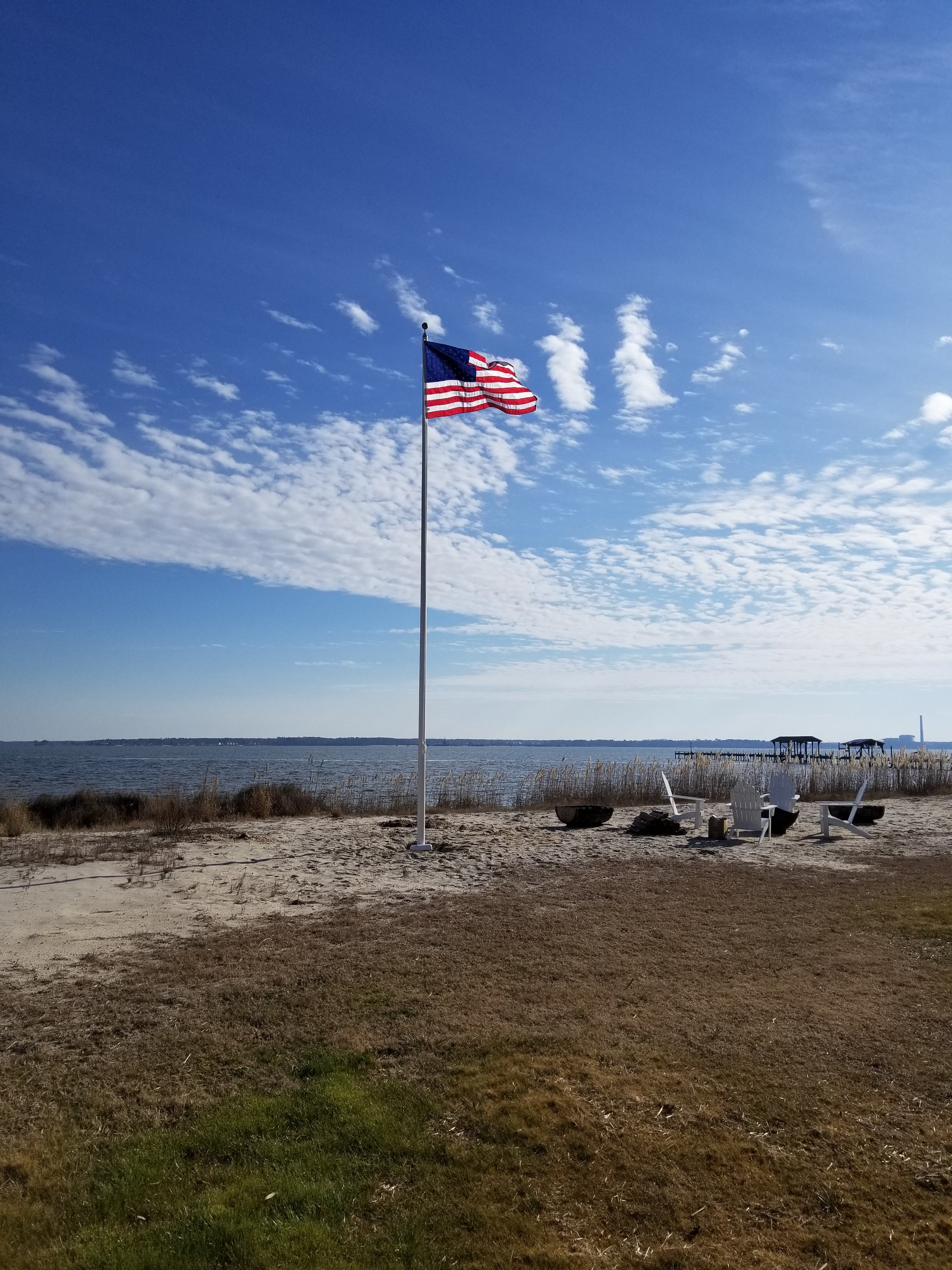 American flag on a pole near a sandy beach with a view of the ocean and a clear blue sky with scattered clouds. White chairs and a fire pit are on the beach.