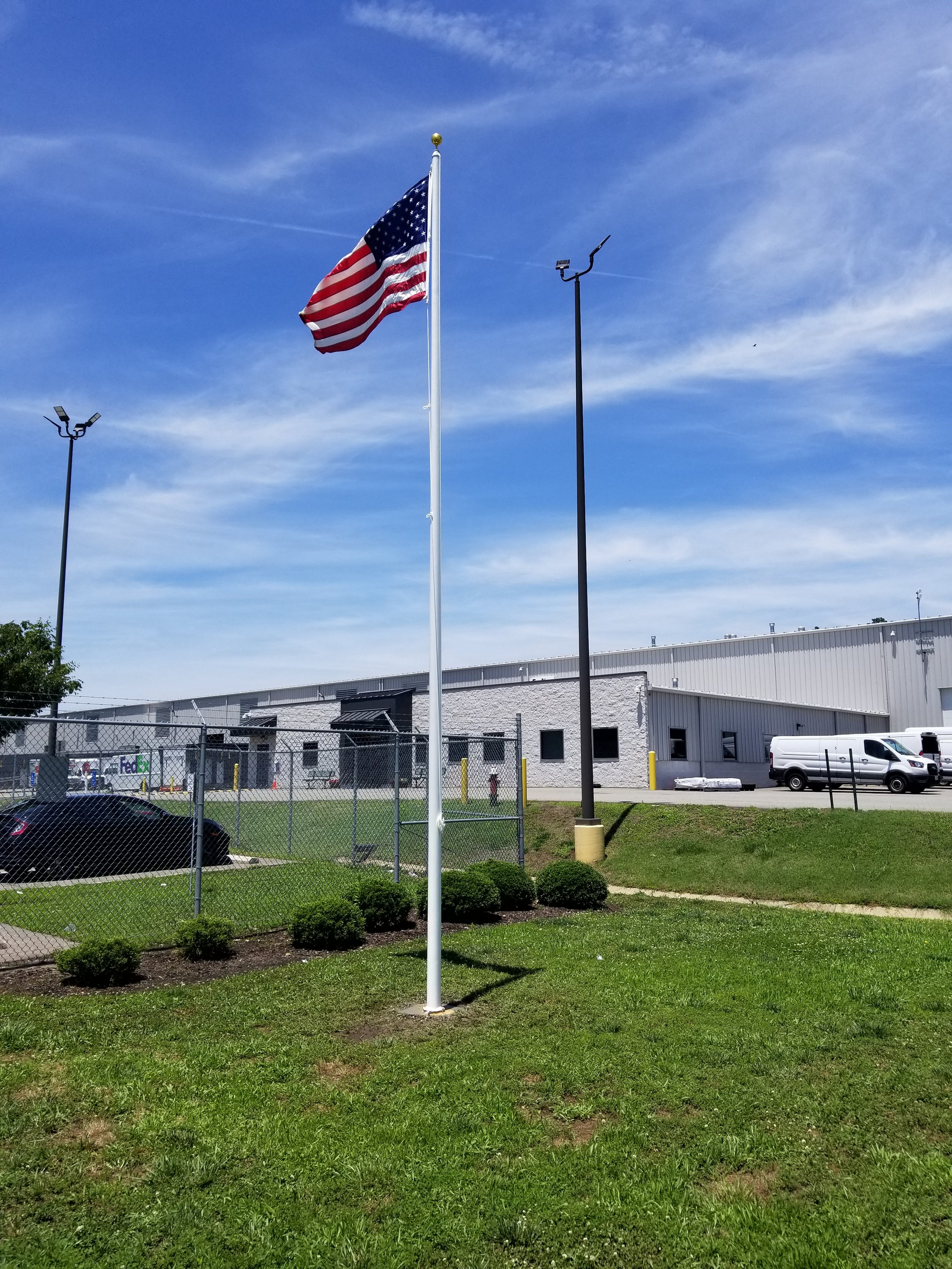 American flag on a pole in front of an industrial warehouse, blue sky background, fenced area, parked vehicles, and greenery.