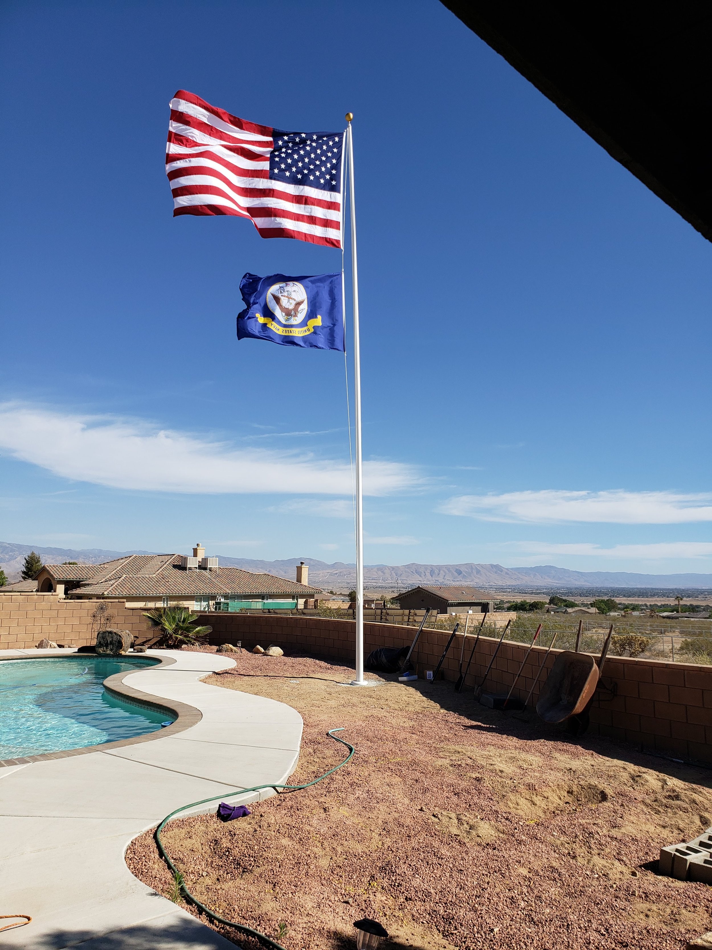 American and U.S. Navy flags flying on a flagpole near a swimming pool in a residential backyard with a view of mountains and clear blue sky.