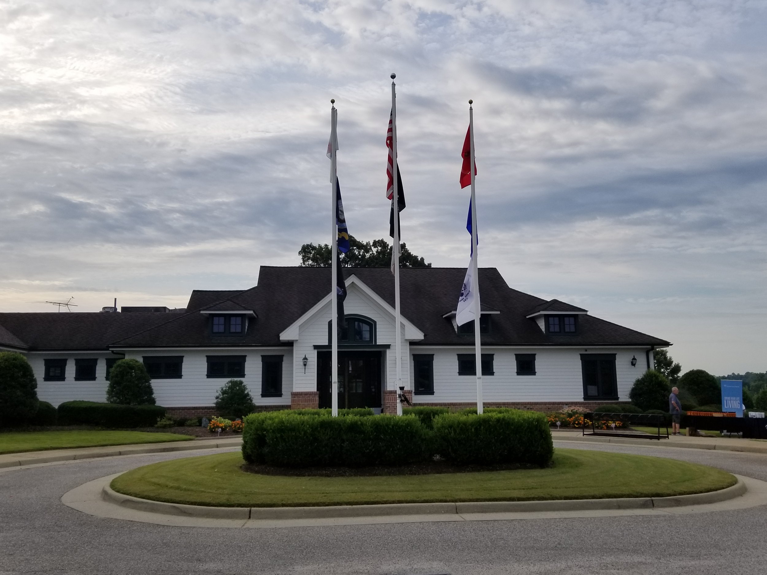 Building with flags in front, circular driveway, and landscaped garden, under cloudy sky.