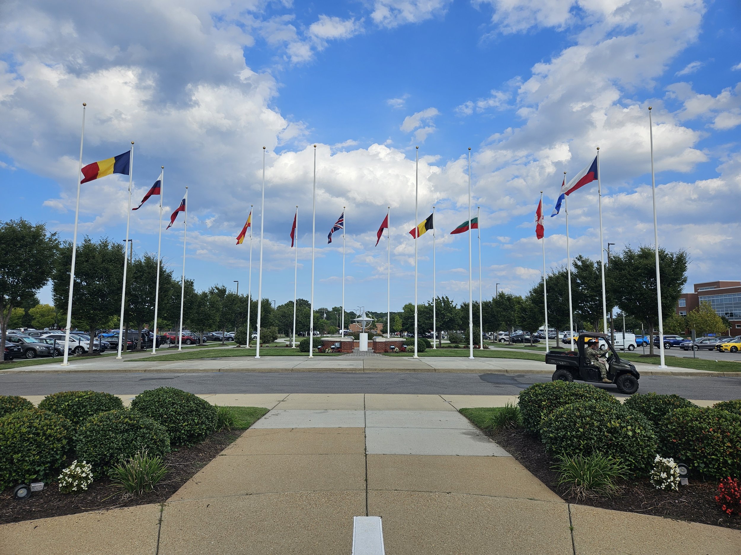 A row of international flags on poles beside a parking lot and landscaped walkway under a partly cloudy sky.