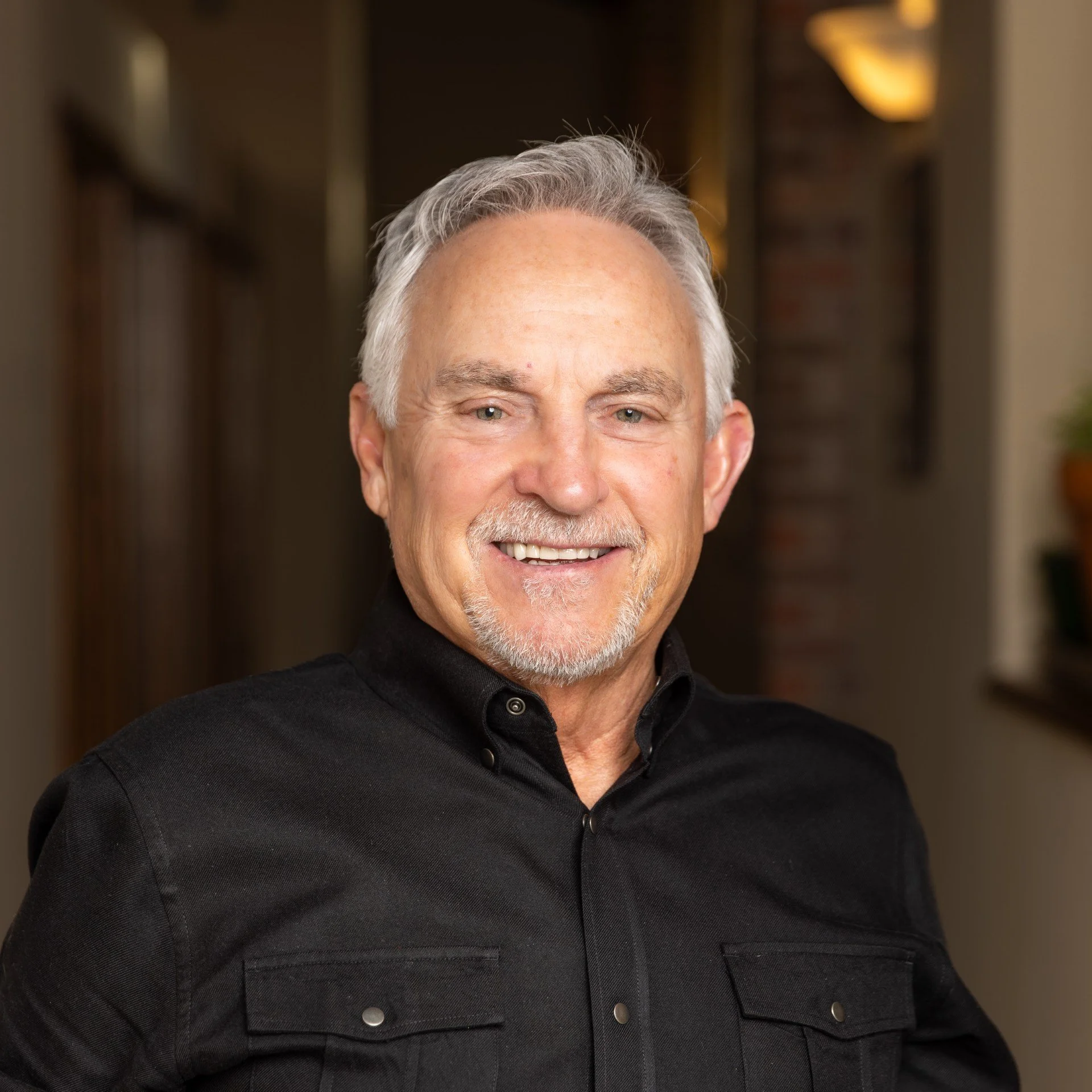 A middle-aged man with gray hair and a beard, wearing a black shirt, smiling indoors in front of a blurred background.