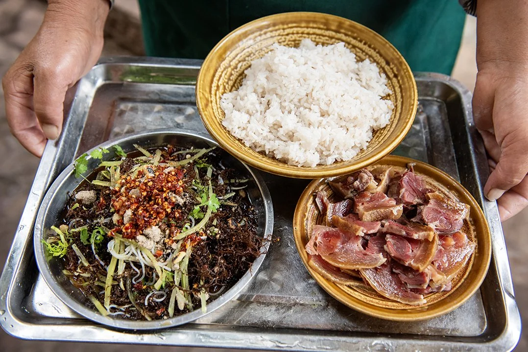 Person holding a tray with three dishes: a bowl of white rice, sliced meat, and a spicy salad with herbs and vegetables.