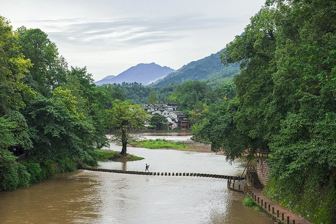 River with stepping stones, surrounded by lush green trees and distant mountains, with a person crossing. Traditional village buildings in the background.
