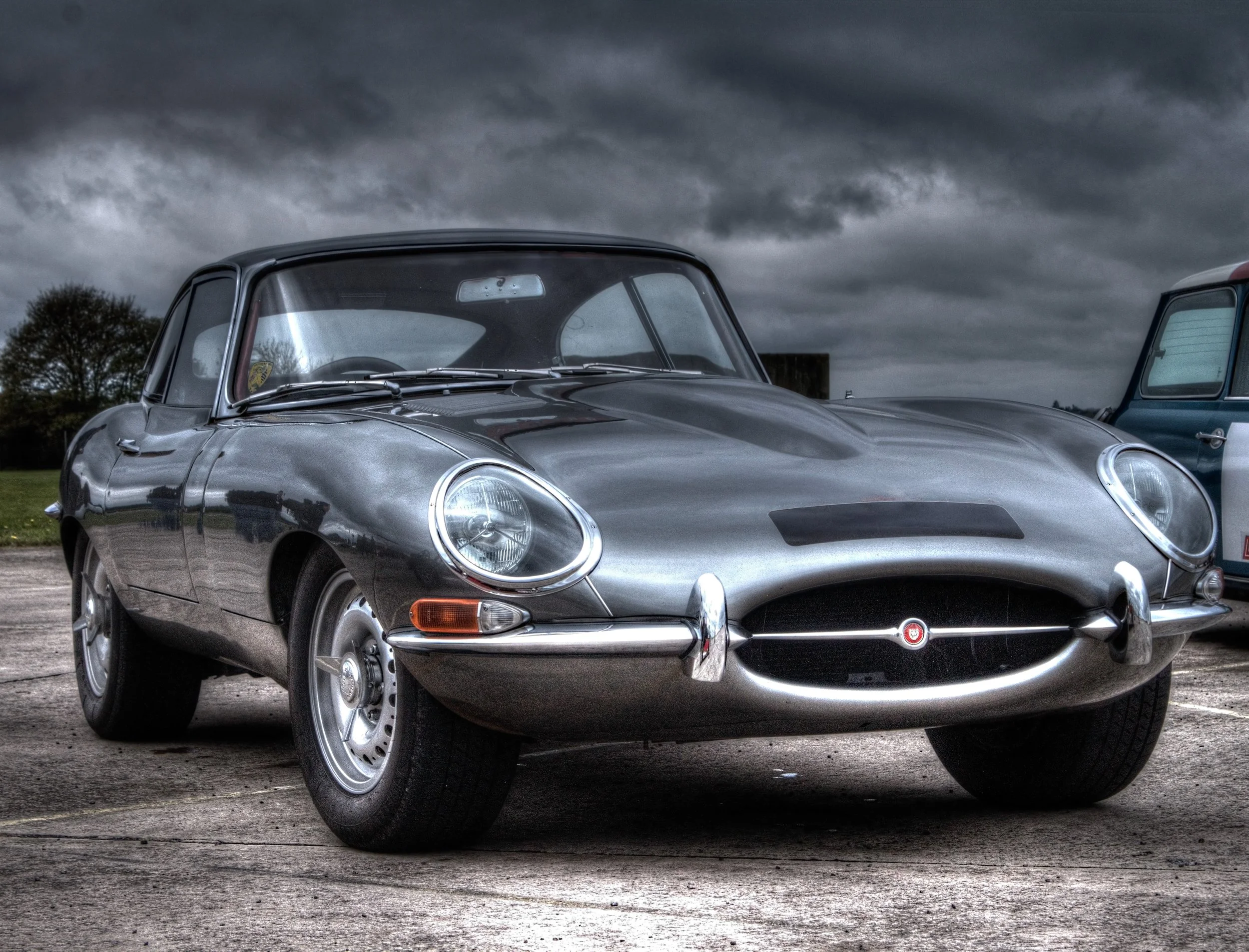 A classic silver Jaguar E-Type sports car parked outdoors under dark clouds.