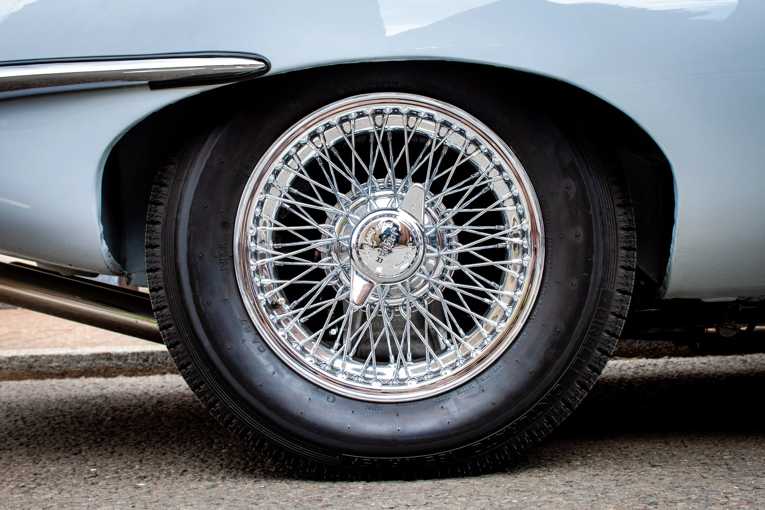 Close-up detail of a Jaguar E-Type car wheel with chrome wire spokes and a black tire, part of a light blue classic car.