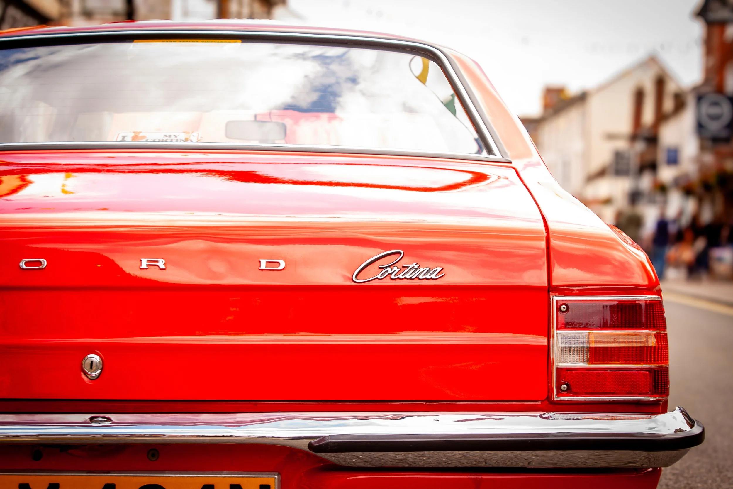 Back view of a red Ford Cortina car parked on a busy street with buildings and people in the background.