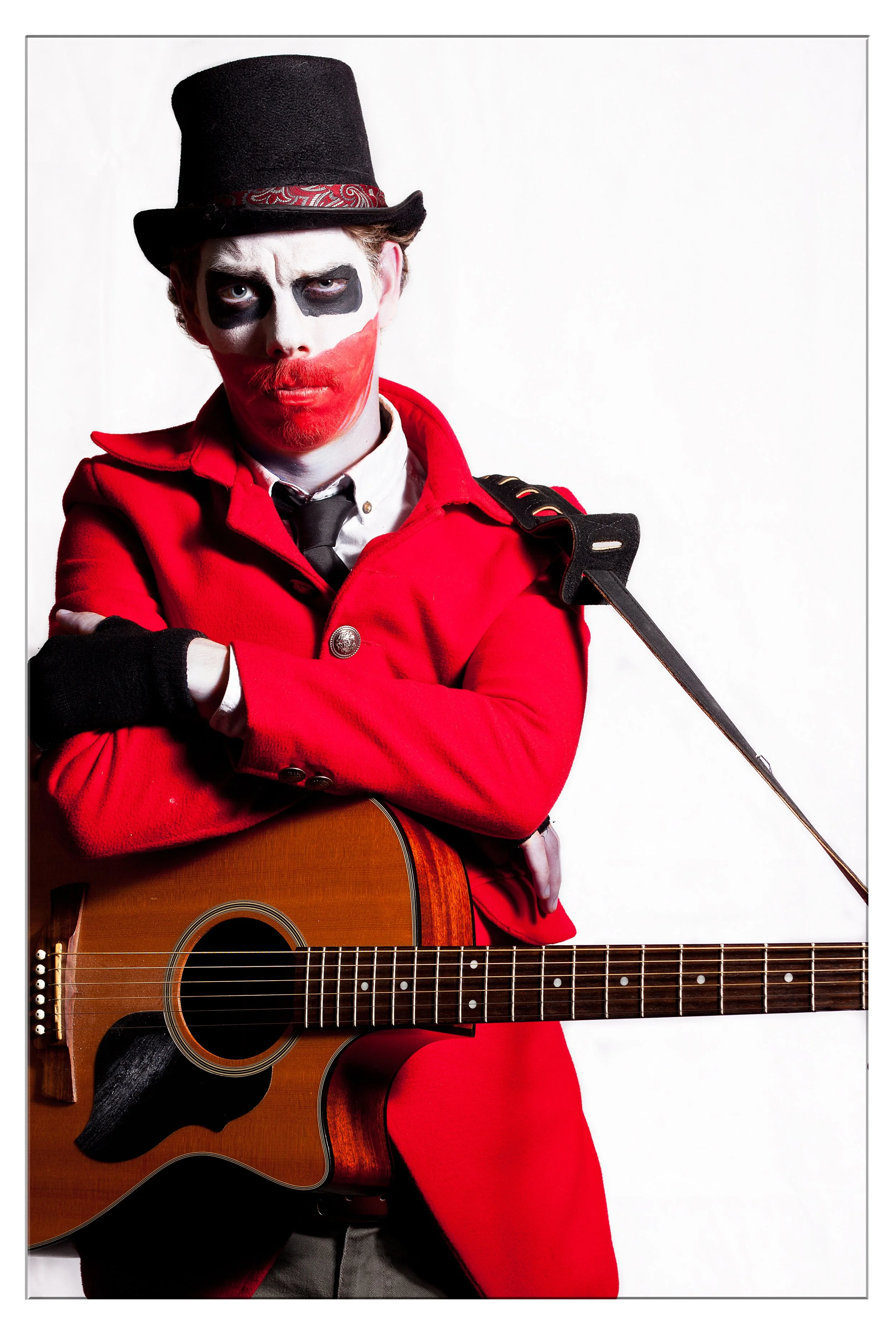 Person dressed as a clown with makeup resembling the Joker, wearing a red coat, black top hat, holding an acoustic guitar, against a plain white background.