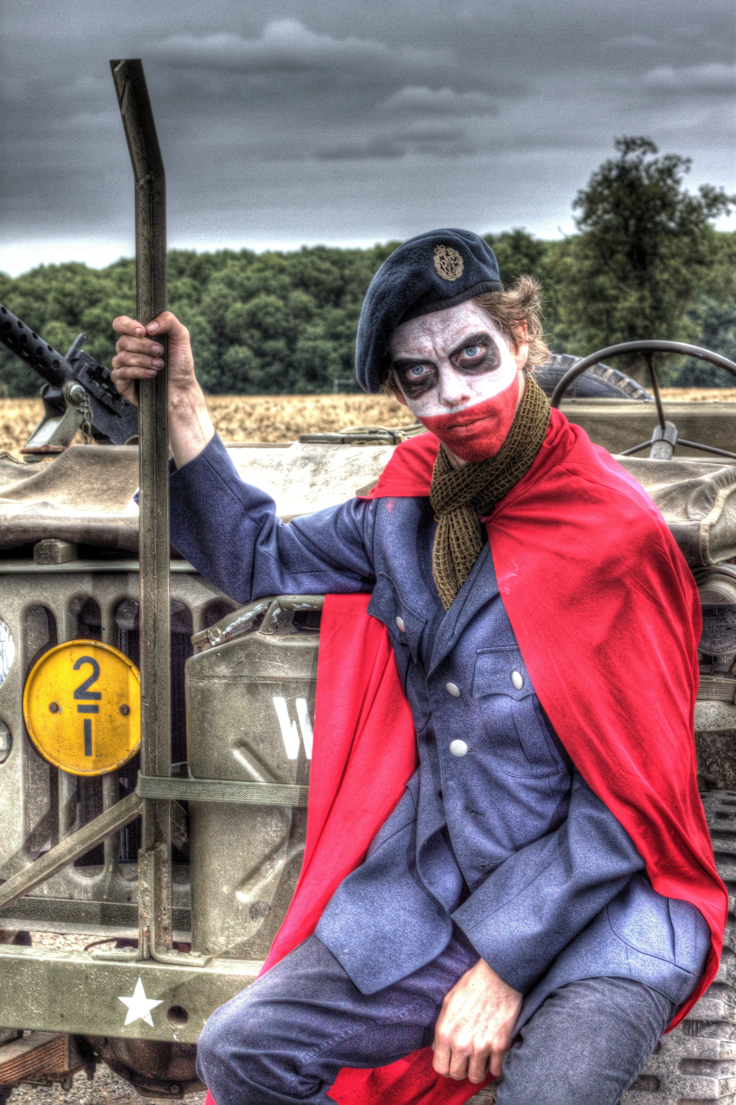 Person dressed as a clown with face paint resembling the Joker, wearing a military beret, a blue uniform, and a red cape, sitting on a military vehicle outdoors on a cloudy day.