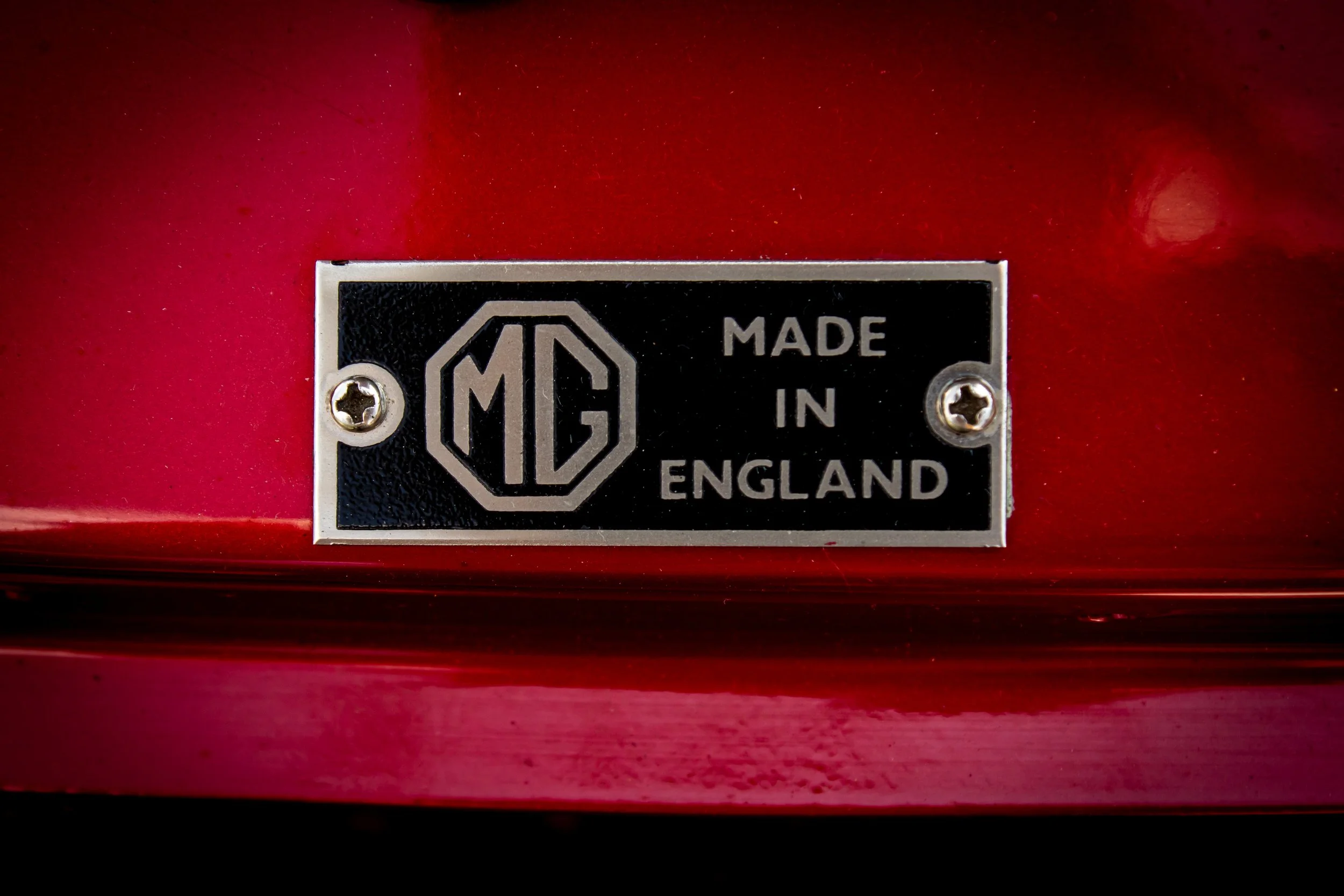 Metal nameplate with MG logo and the words 'Made in England' attached to a red surface.