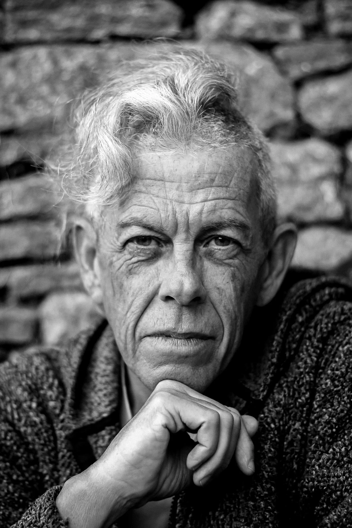 Black-and-white portrait of an caucasian man with gray hair, intense eyes, and a contemplative expression, resting his chin on his hand in front of a stone wall.