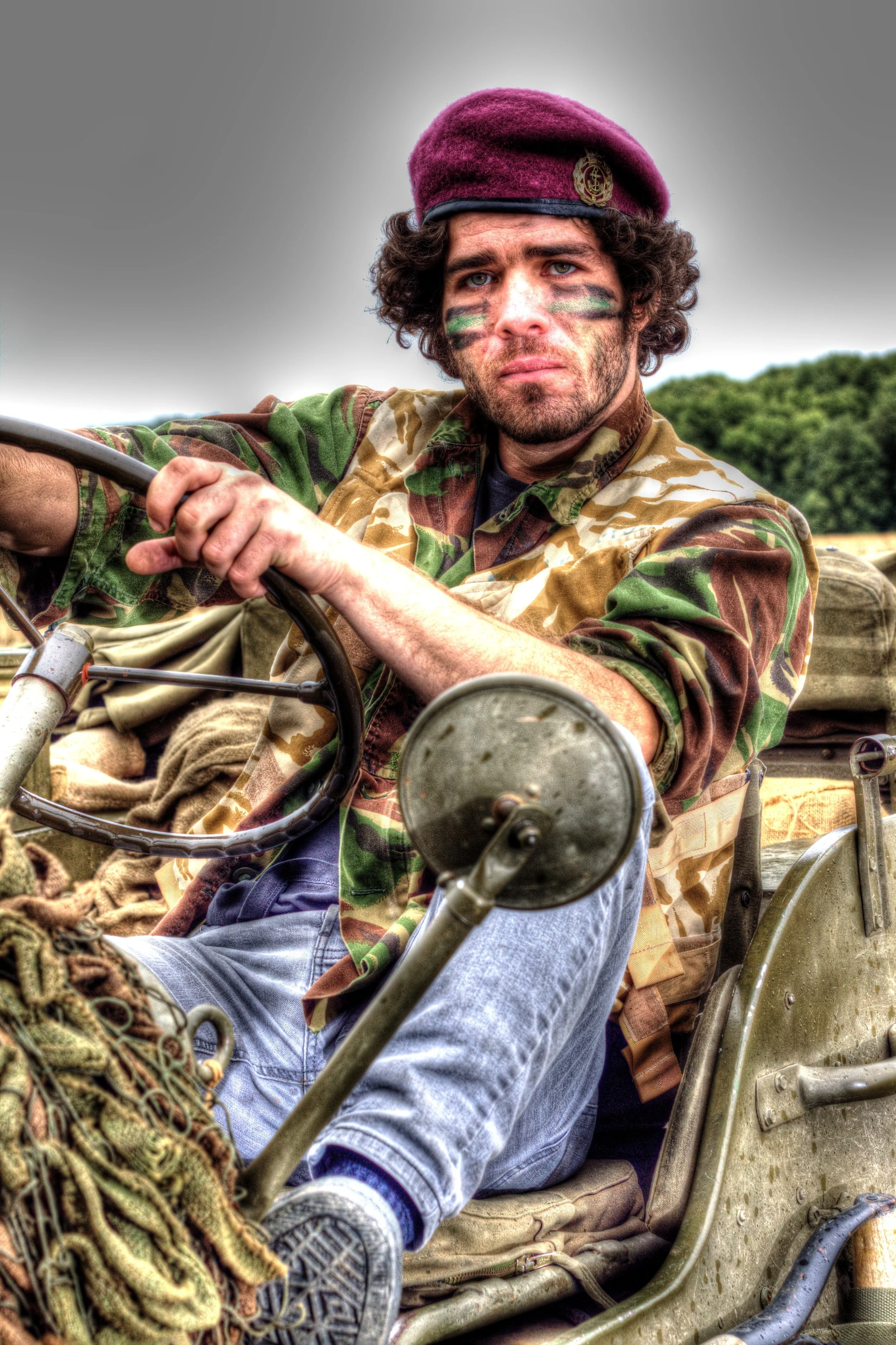 Man in military camouflage uniform with face paint, sitting on a vintage military vehicle, wearing a maroon beret, holding the vehicle's steering wheel, outdoors with a cloudy sky and trees in the background.