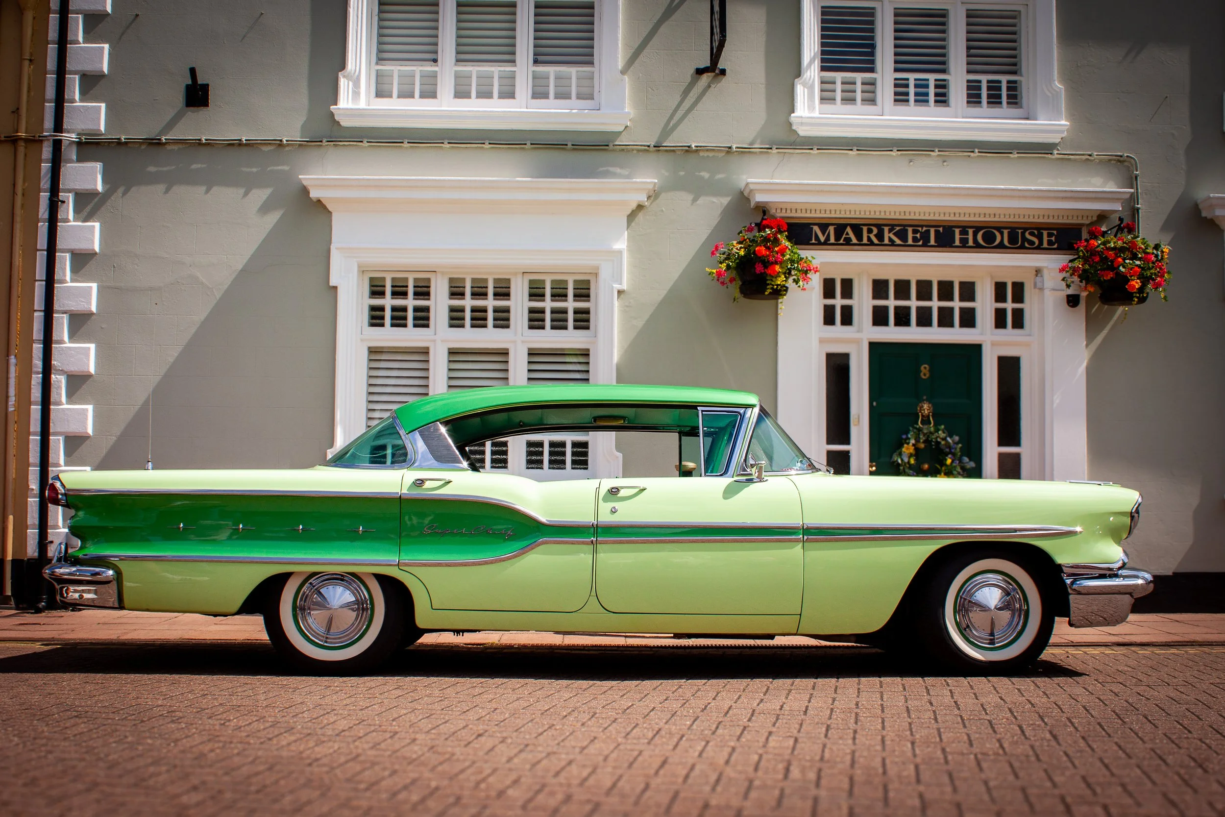 A vintage green and cream-colored classic car parked in front of a building with white window shutters and flower baskets, labeled 'Market House' above the door.