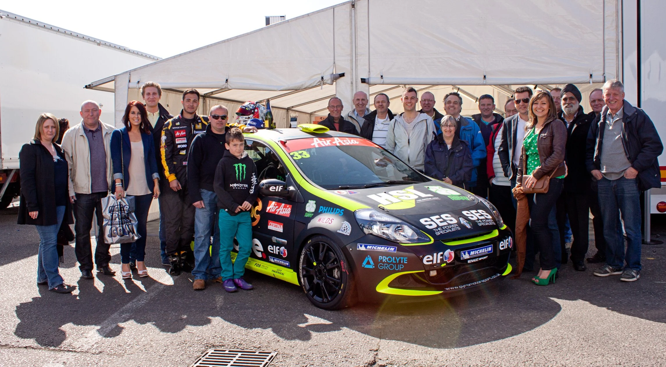 A group of people, including a young boy, surrounding a black racing car with green accents, at a motorsport event. The group is standing outdoors under a tent, with a trailer in the background.