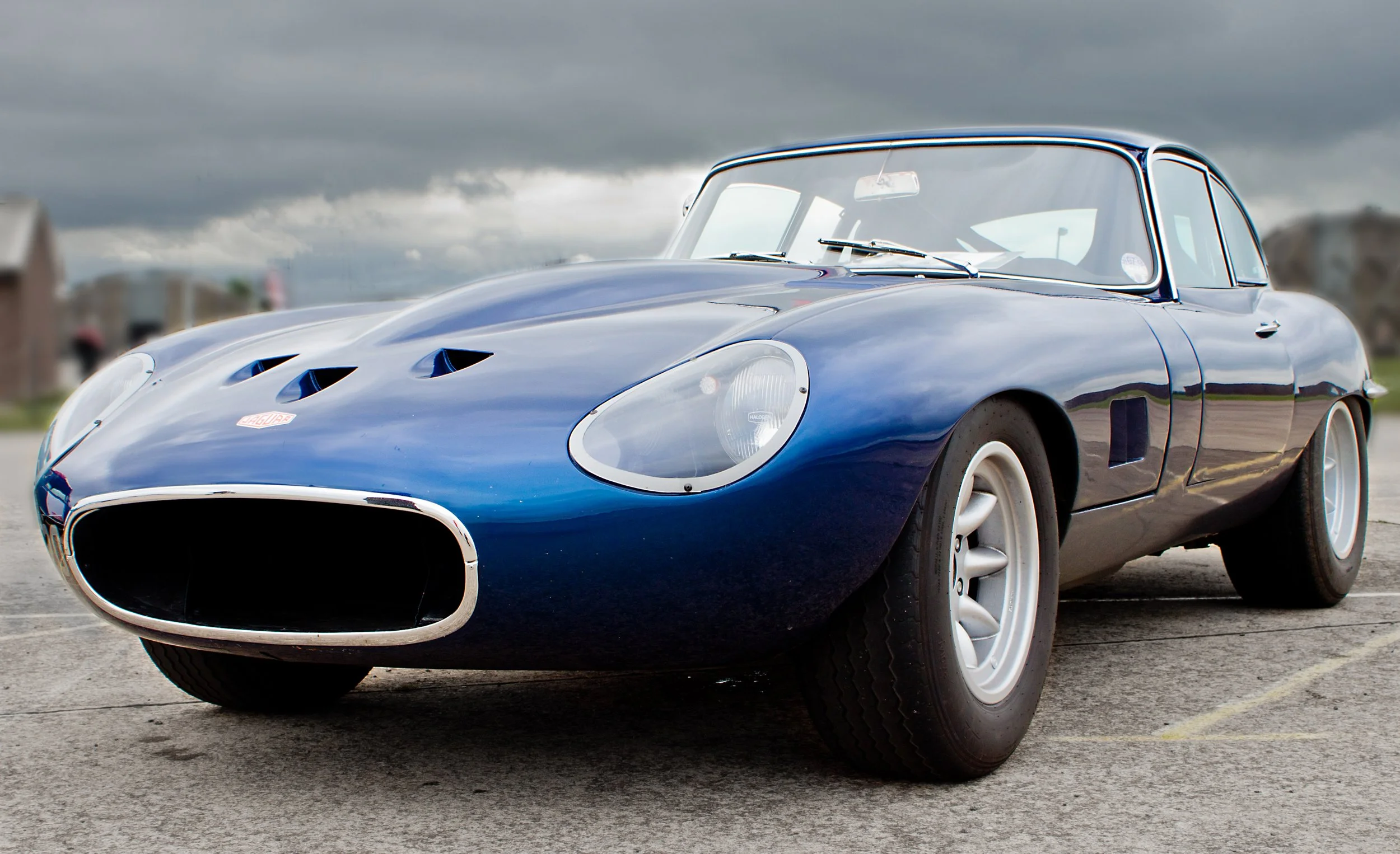 A classic blue Jaguar E-Type sports car parked outdoors on a cloudy day.