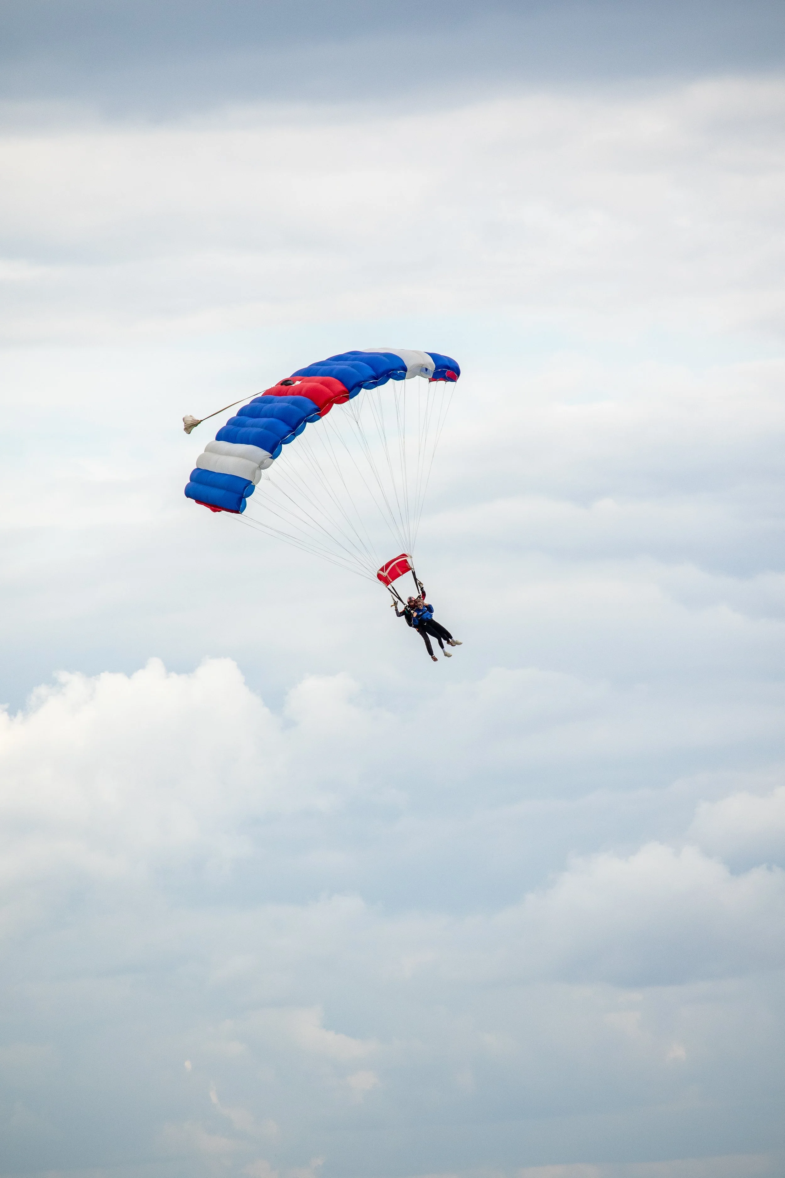 Two people parasailing with a blue, white, and red parachute in a cloudy sky.