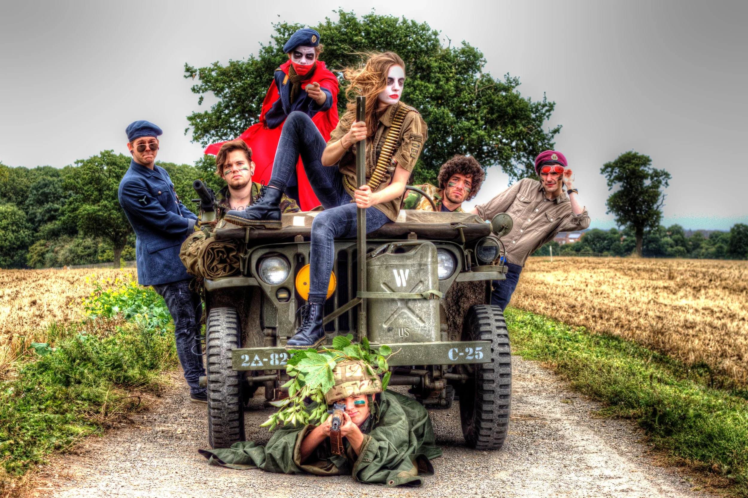 Group of six people dressed in military and costume attire, positioned on and around a military jeep in a countryside setting with trees and open fields.