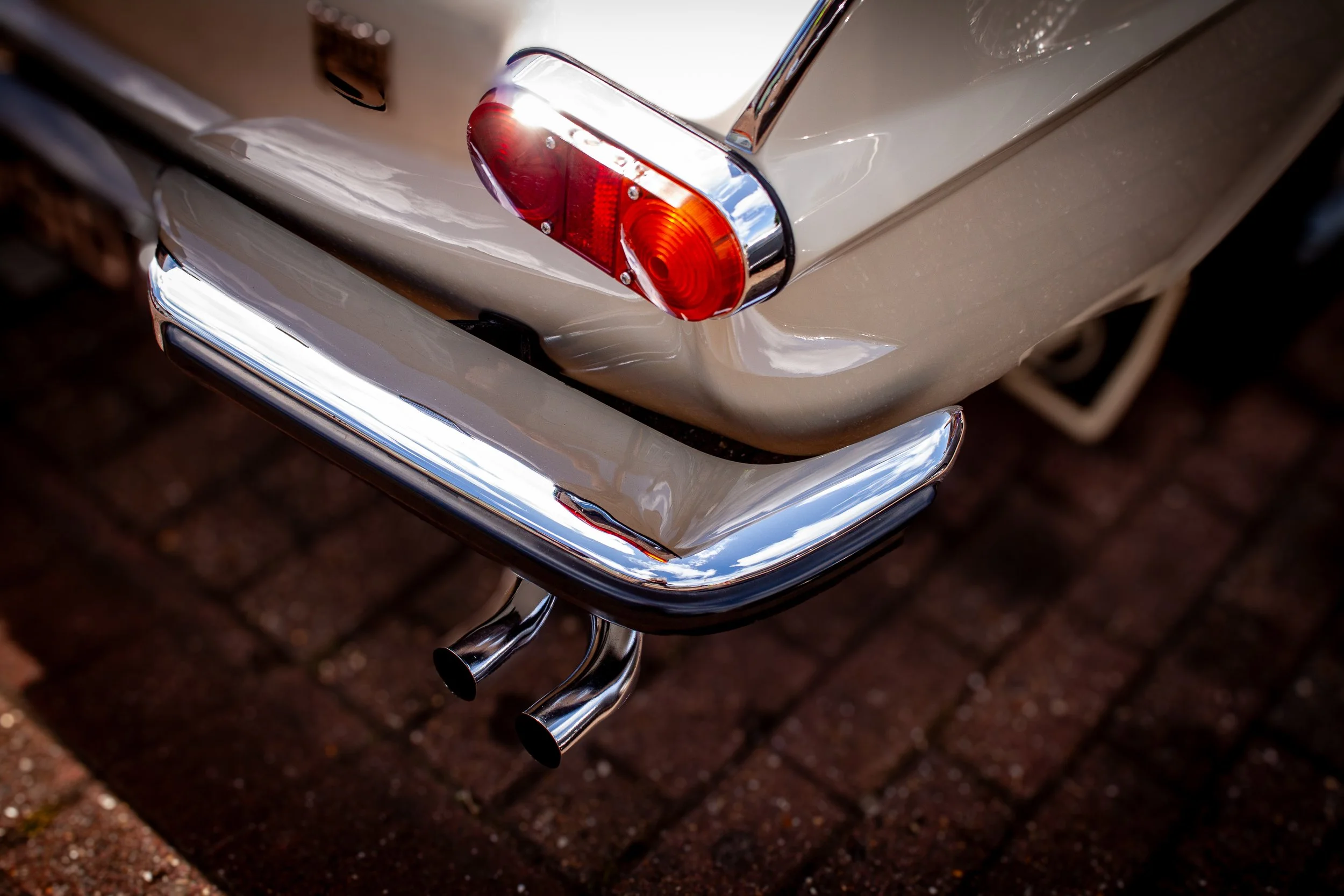 Close-up of the rear end of a beige vintage car with chrome accents, showing dual exhaust pipes, red tail light, and part of the bumper, parked on brick pavement.