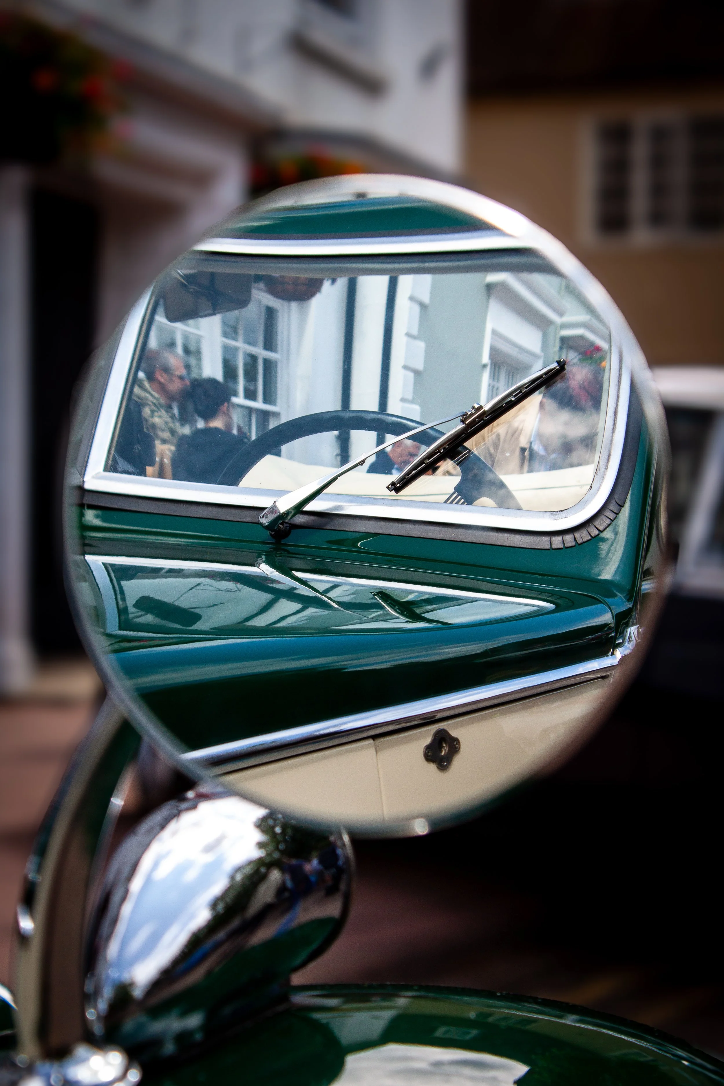 View of a vintage green car reflected in a convex side mirror, showing a window and people sitting inside the building.