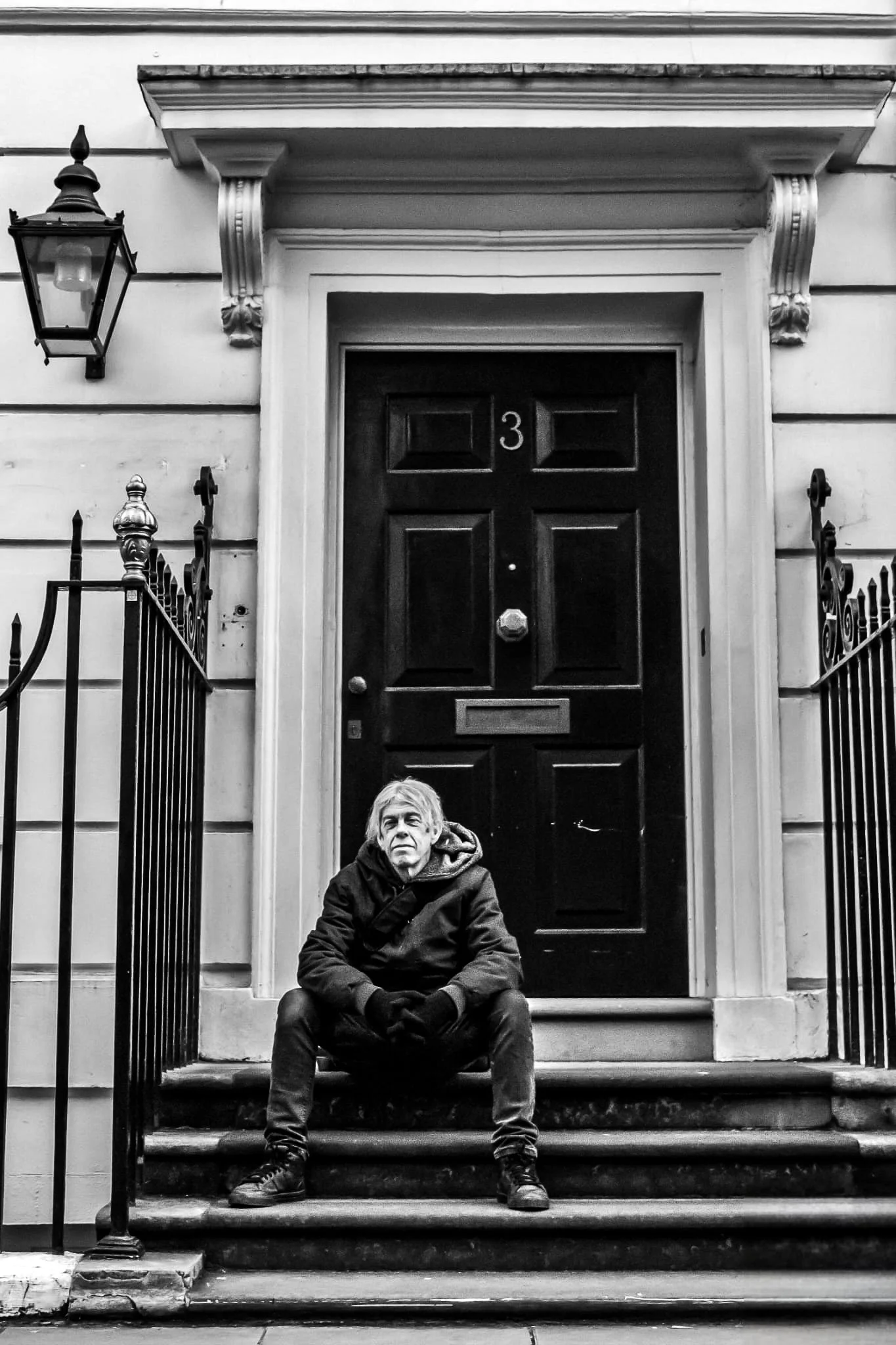 A man sitting on the steps in front of a black door with a house number 3, flanked by decorative railings and a wall-mounted lantern.
