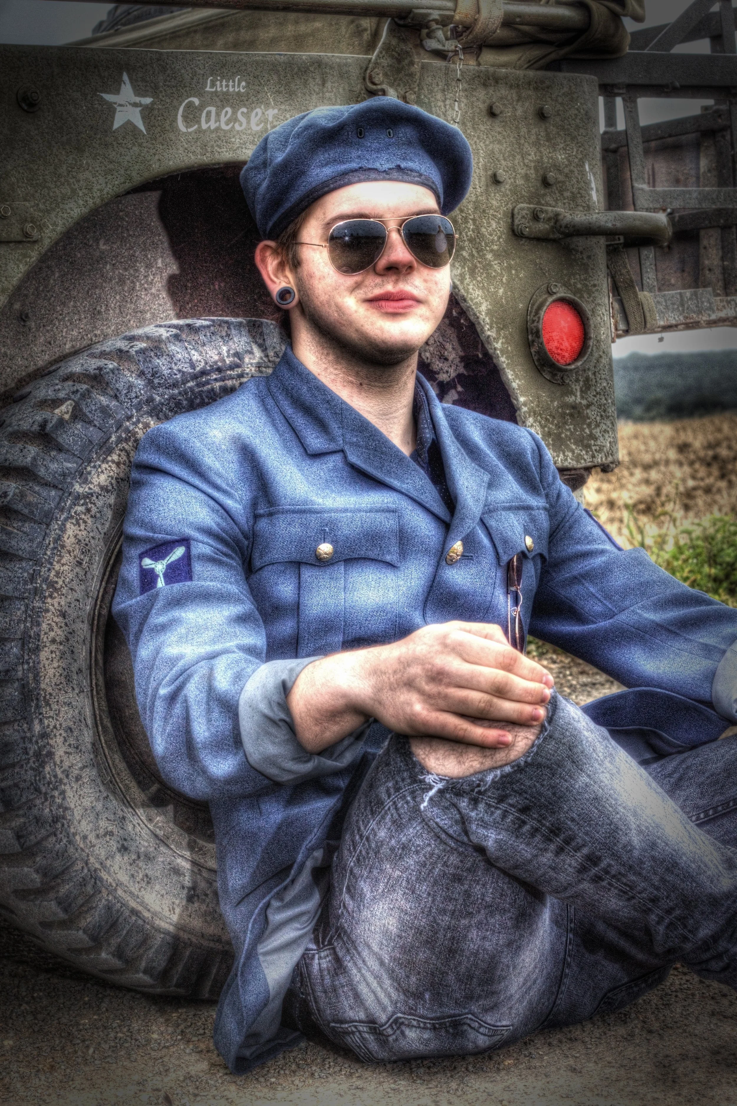 Young man wearing sunglasses, a blue military-style jacket, and a blue beret, sitting on the ground and leaning against a military vehicle labeled "Little Caesar" with a star emblem. The background shows an open field.