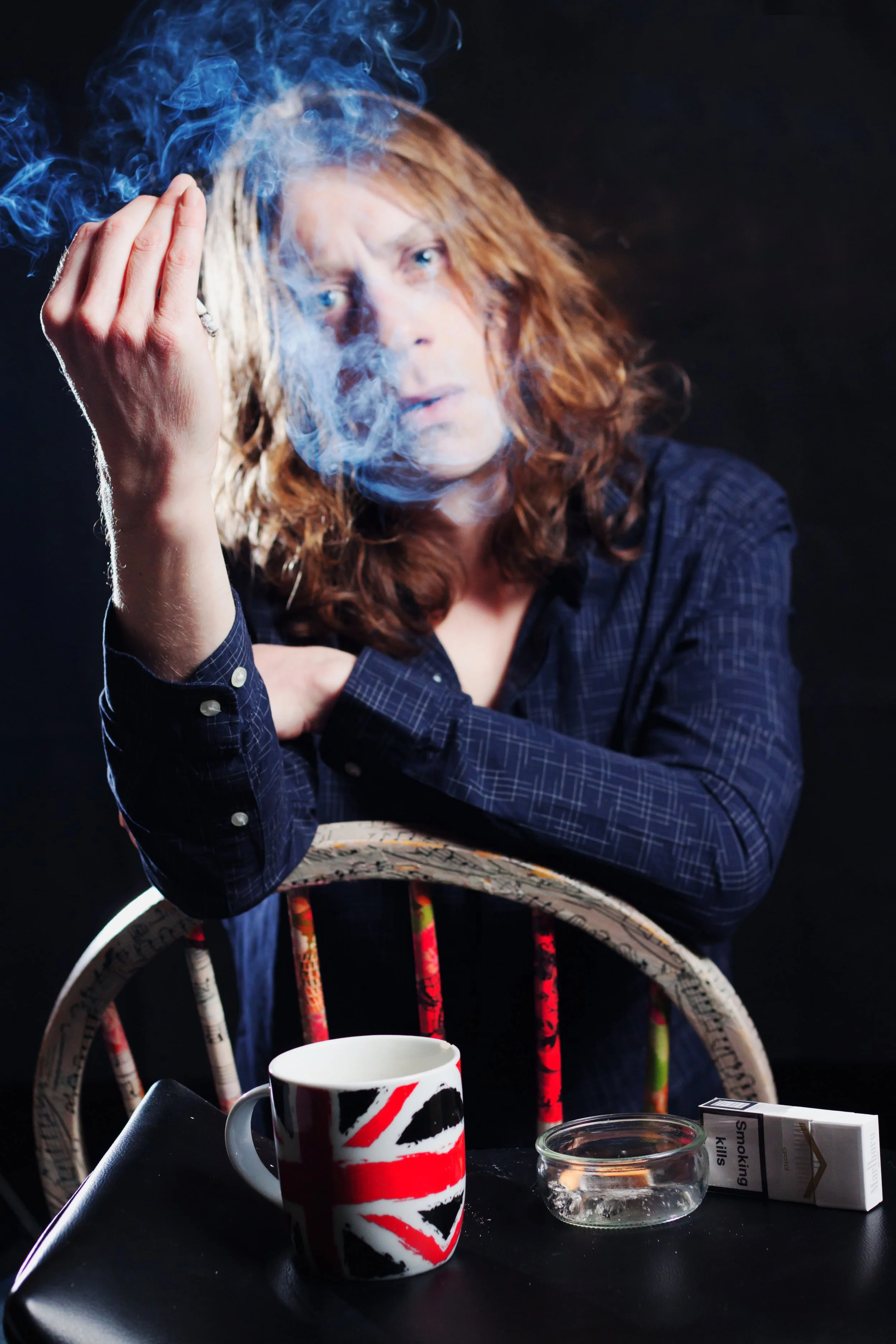 A woman with red hair sitting at a table, smoking a cigarette, with smoke around her face. On the table, there is a ceramic mug with a Union Jack design, a small glass ashtray, and a pack of cigarettes labeled "Smoking Kills."