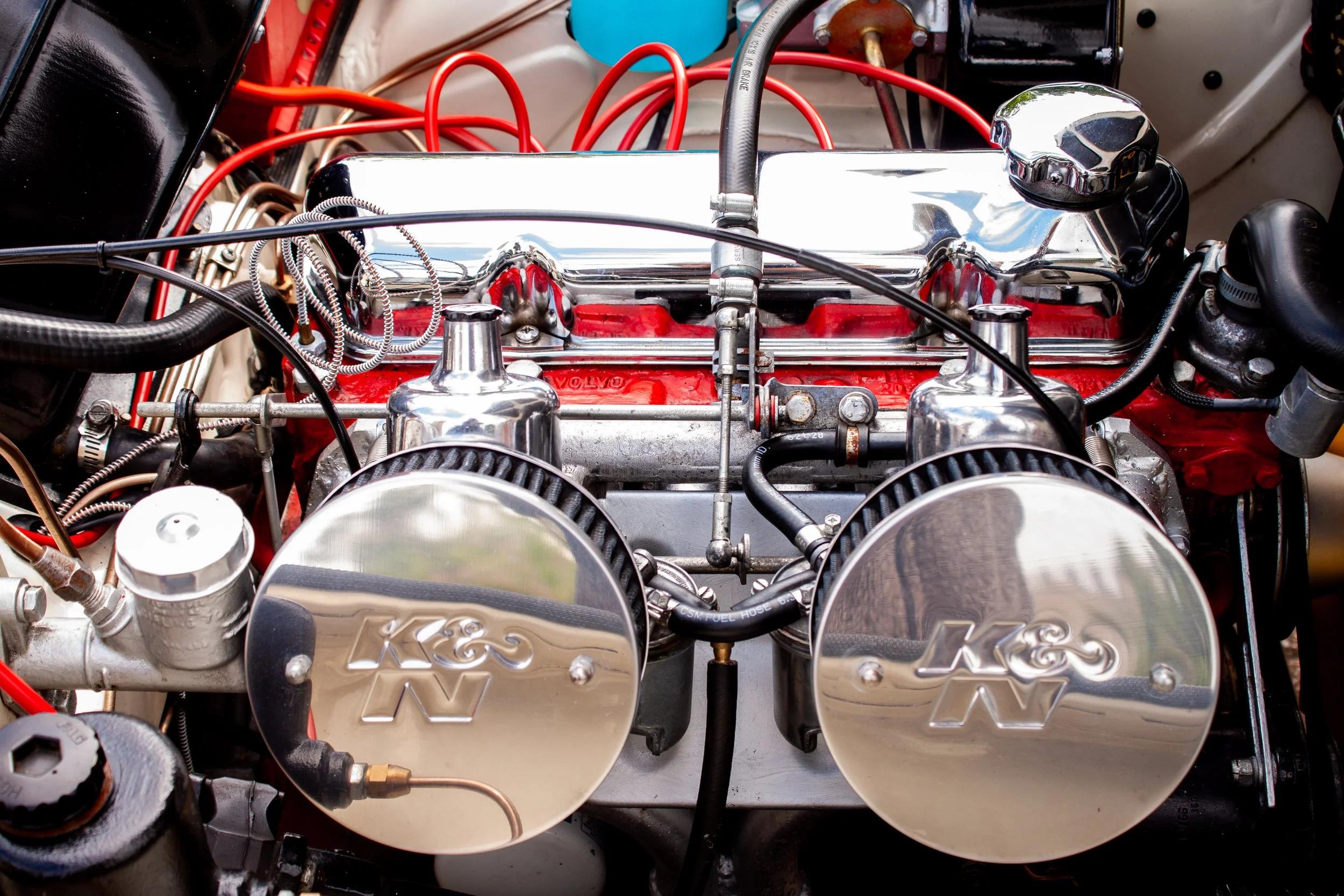 Close-up of a vintage car engine featuring dual chrome air filters labeled K&N, a red valve cover, and various wires and hoses.