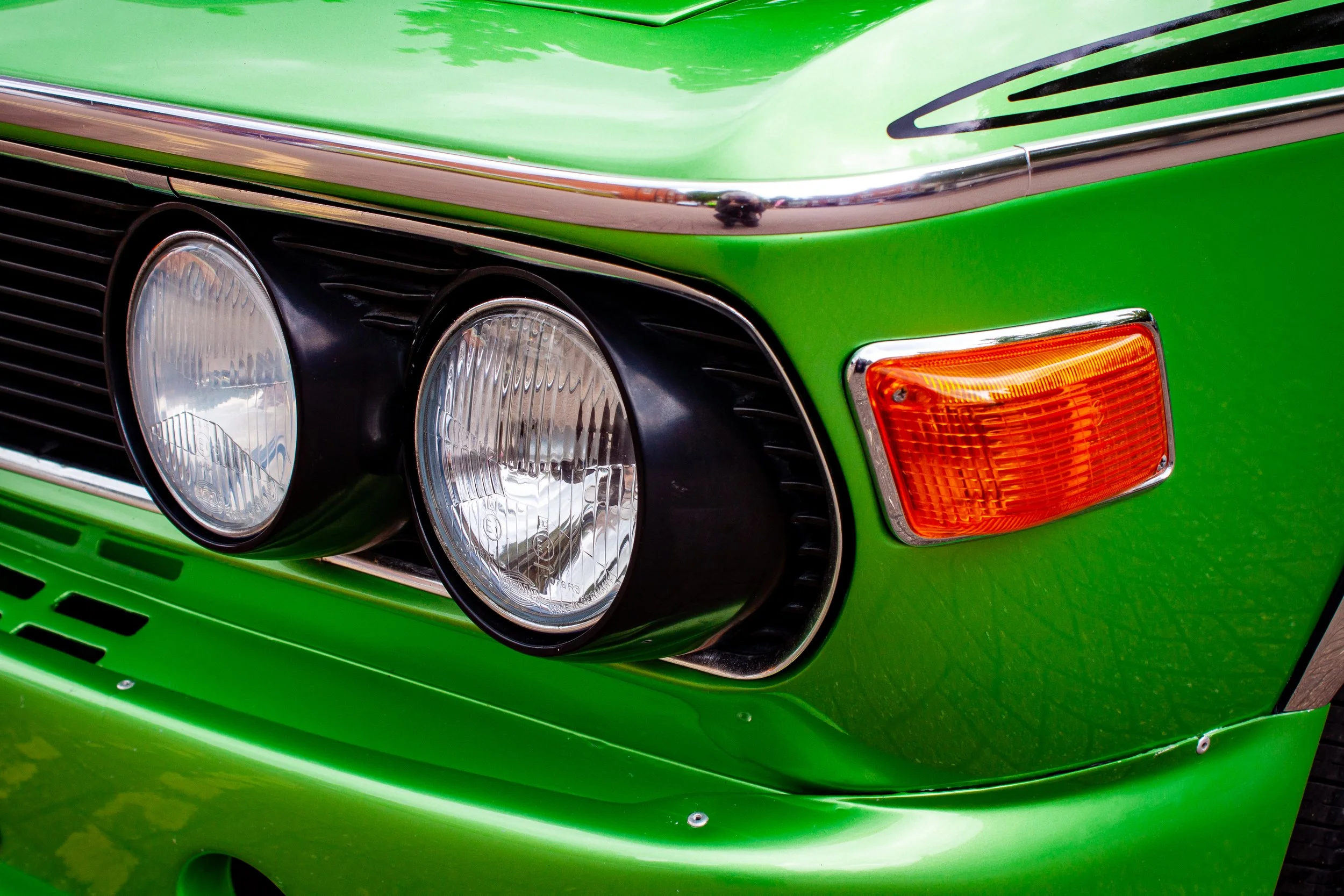 Close-up of the front of a bright green vintage car with dual round headlights, orange turn signals, and chrome accents.