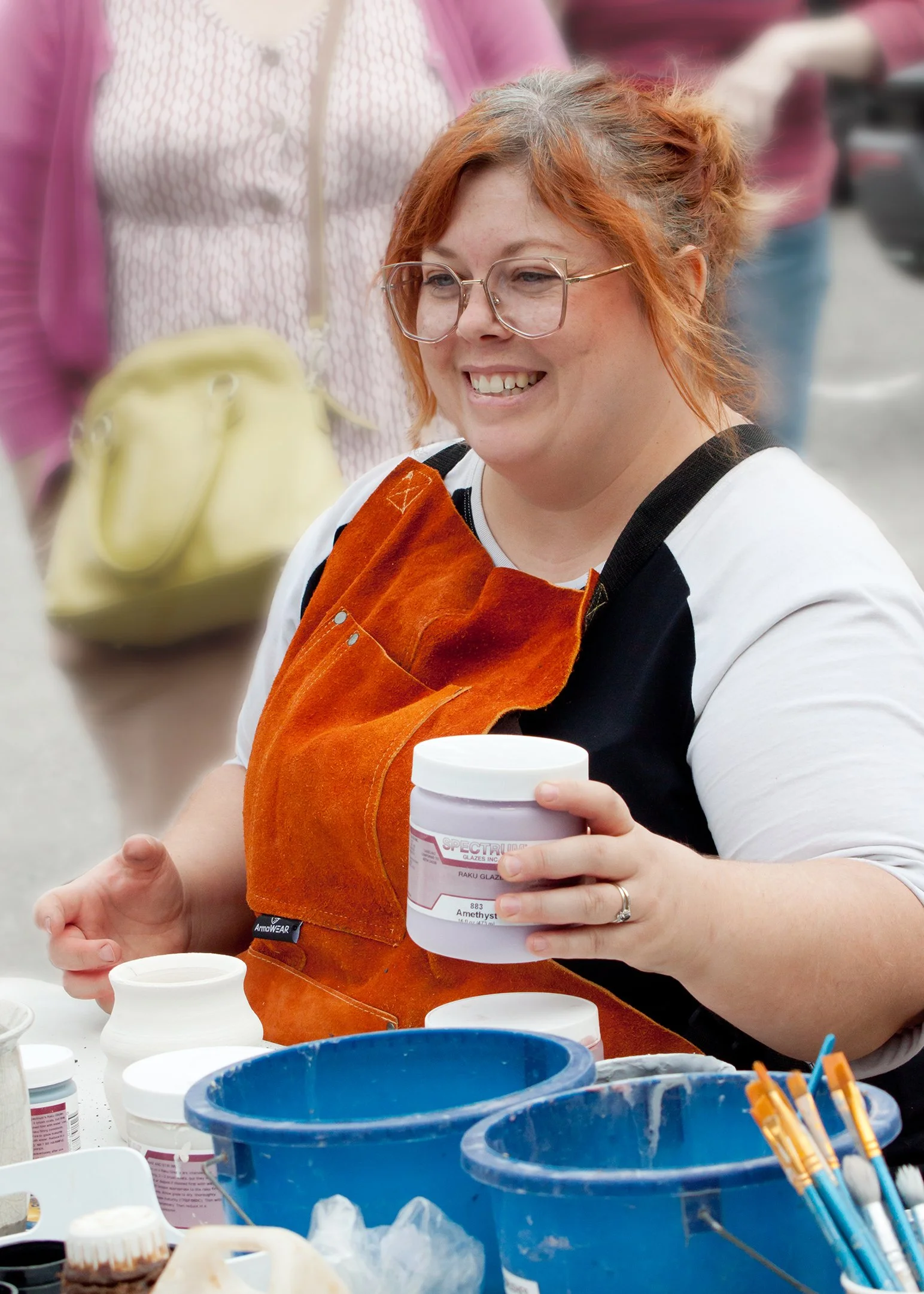 A woman with red hair, glasses, and a white and black shirt, smiling while holding a jar of amethyst-colored powder during a craft or art project at a table with various containers and tools.