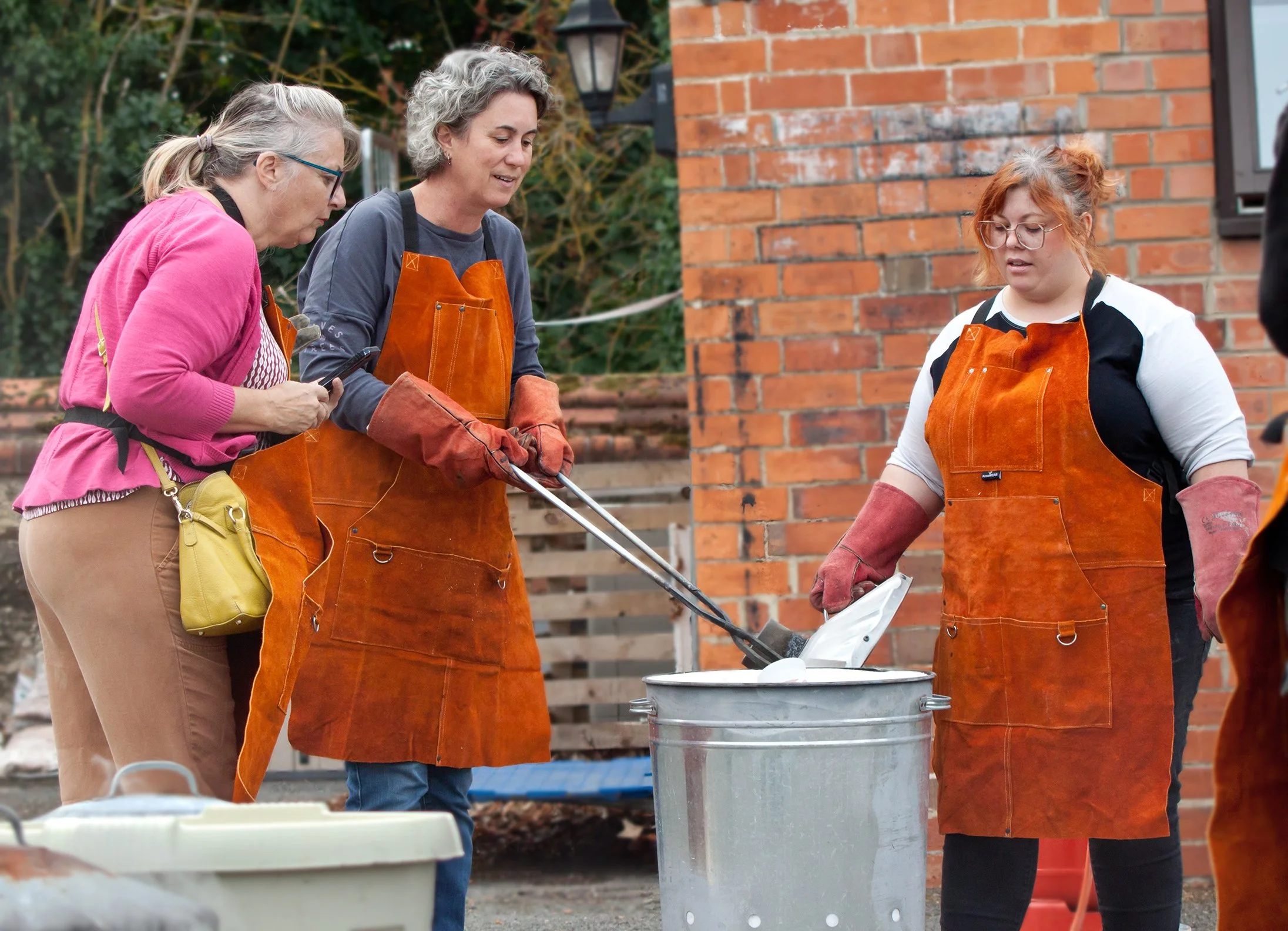 Three women wearing orange aprons and gloves outdoors near a brick wall, cooking food on a large silver pot with an outdoor setting visible in the background.