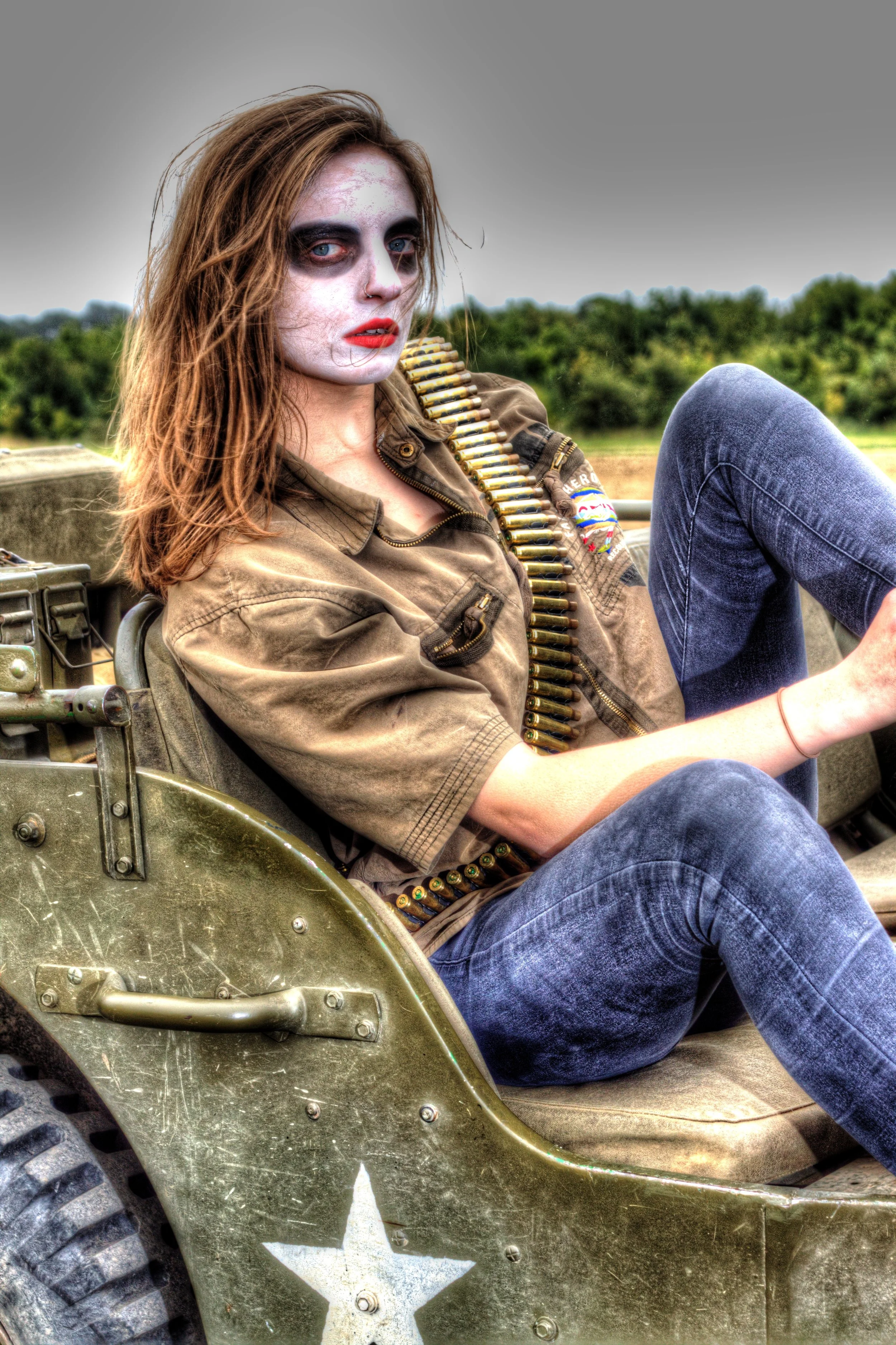 A woman with face makeup resembling a zombie sits in a military vehicle with a large star emblem, wearing a brown shirt and a bandolier of ammunition.