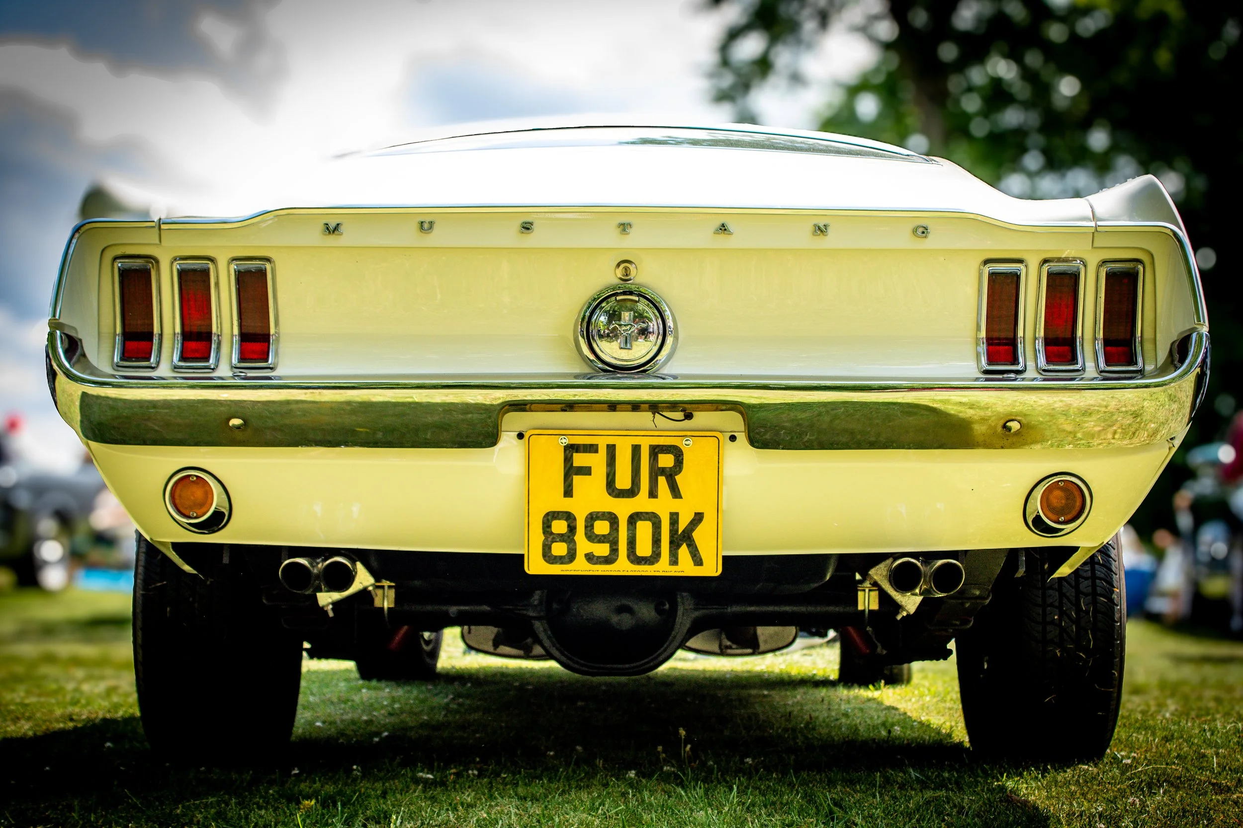 Rear view of a vintage yellow Ford Mustang car with a license plate reading 'FUR 890K', parked on grass during daytime.