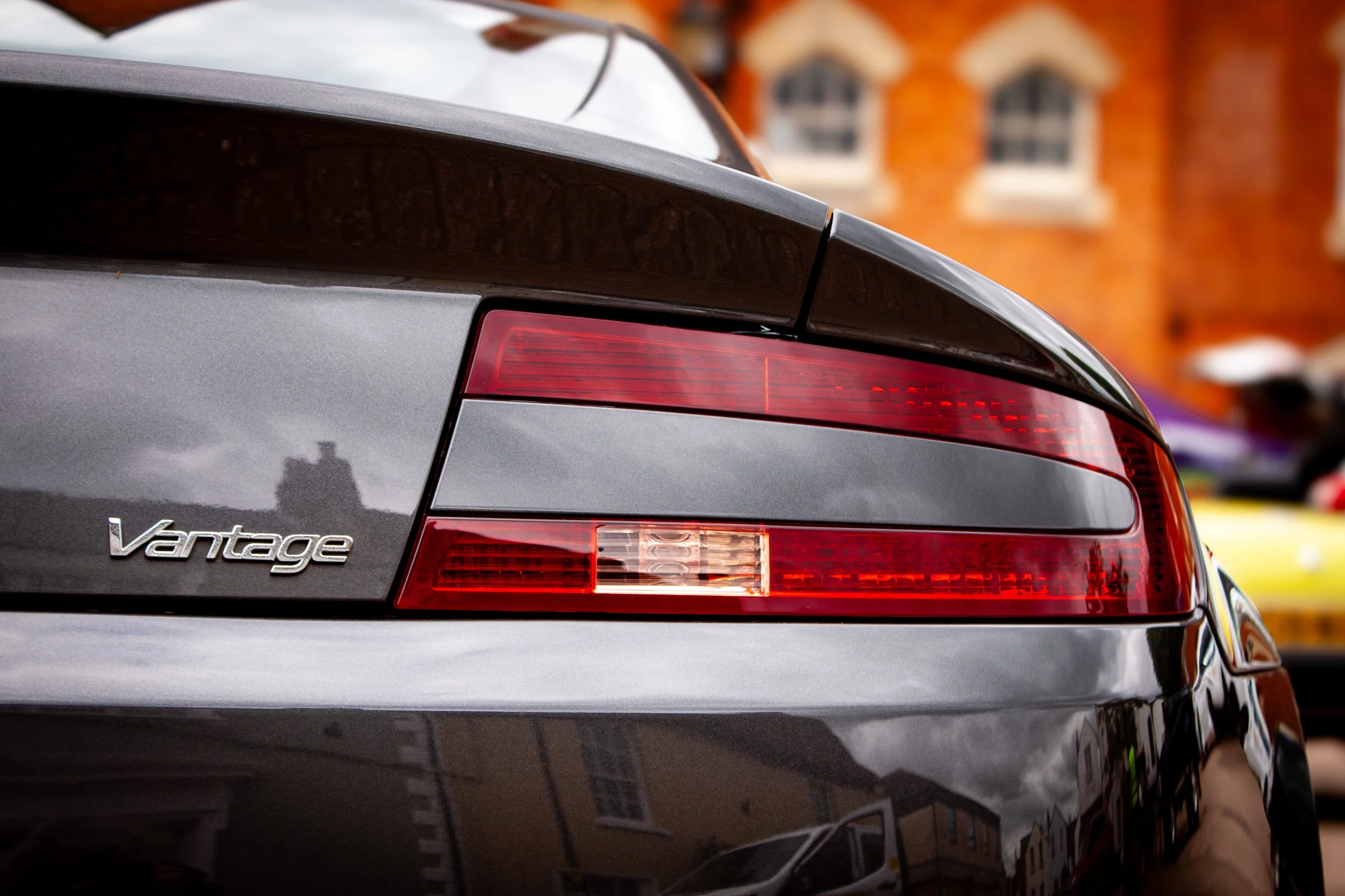 Close-up detail of the rear of a grey Aston Martin Vantage car, showing the taillight and a chrome badge indicating the model.