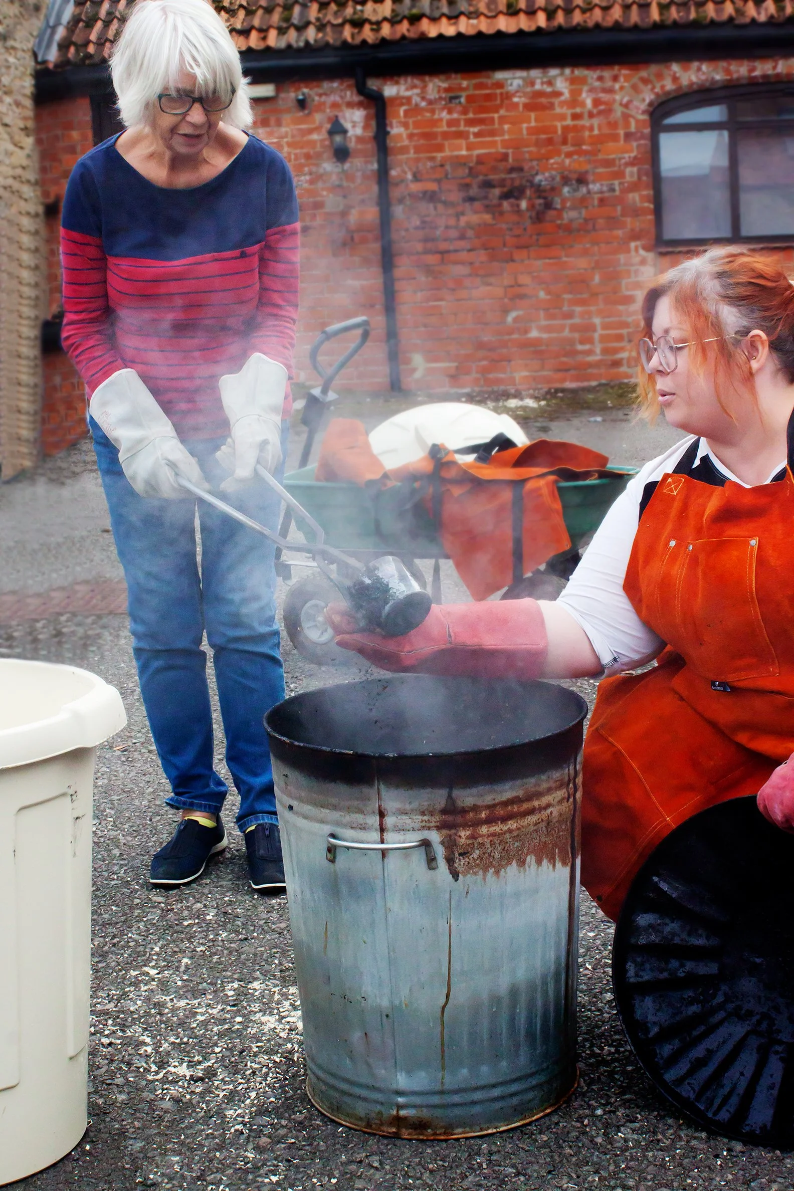 Two women smoking meat over a barrel grill outdoors, one standing and one sitting, using tongs, with a brick building in the background.