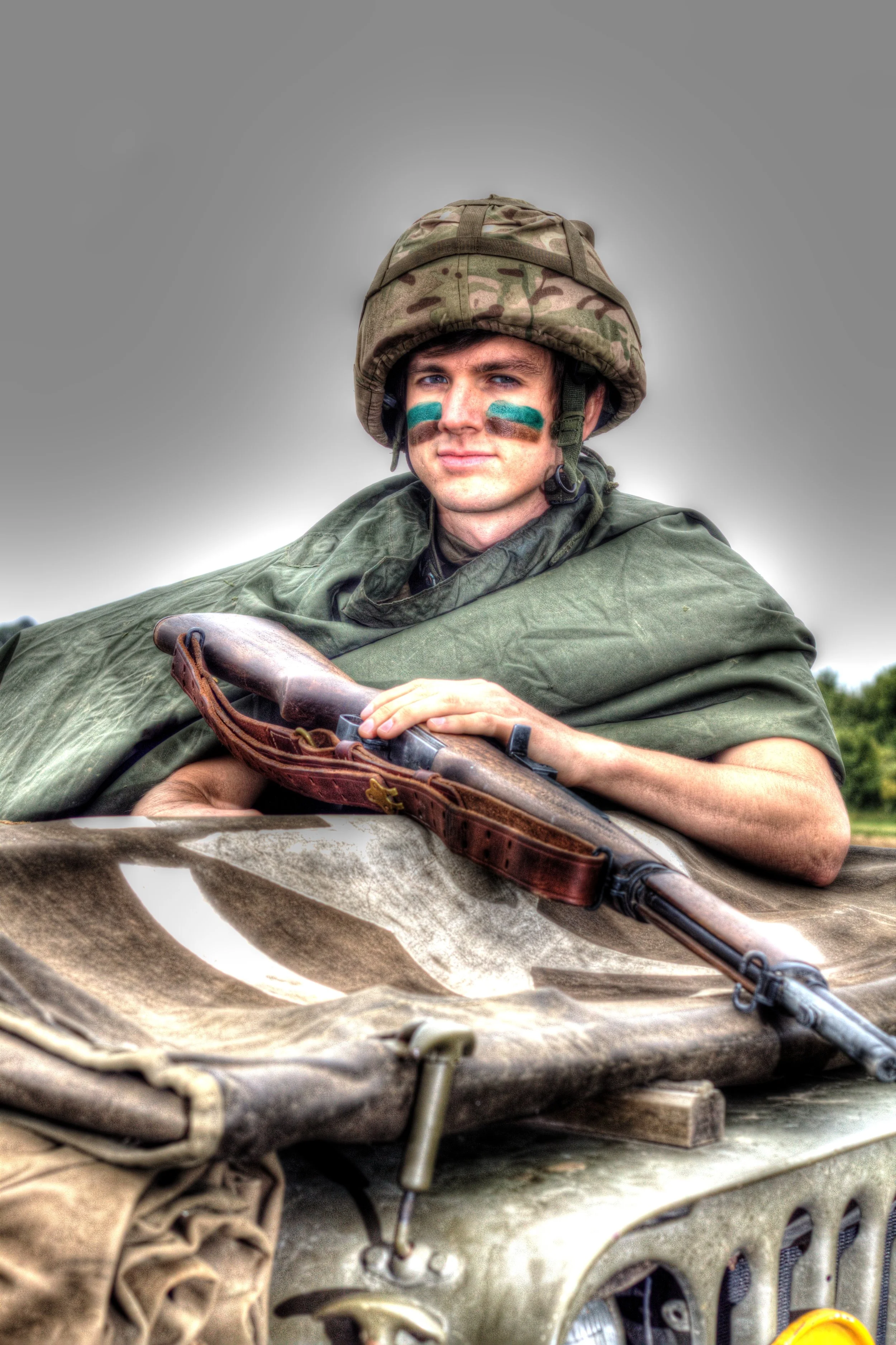 A soldier in camouflage uniform and face paint, wearing a helmet, holding a rifle, and sitting on the front of a military vehicle outdoors.