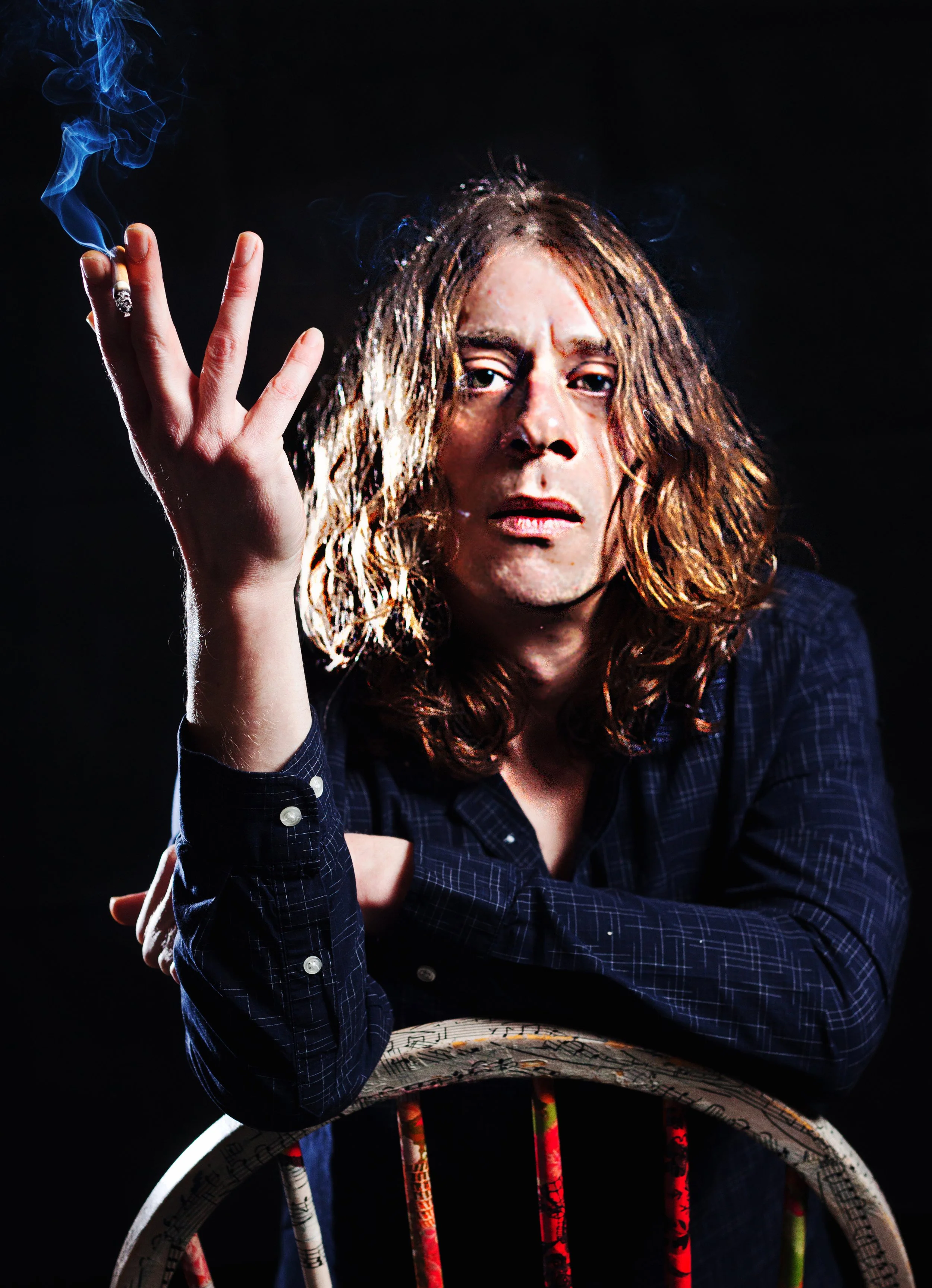 A person with long, wavy hair holding a lit cigarette with smoke rising, looking serious, against a dark background.