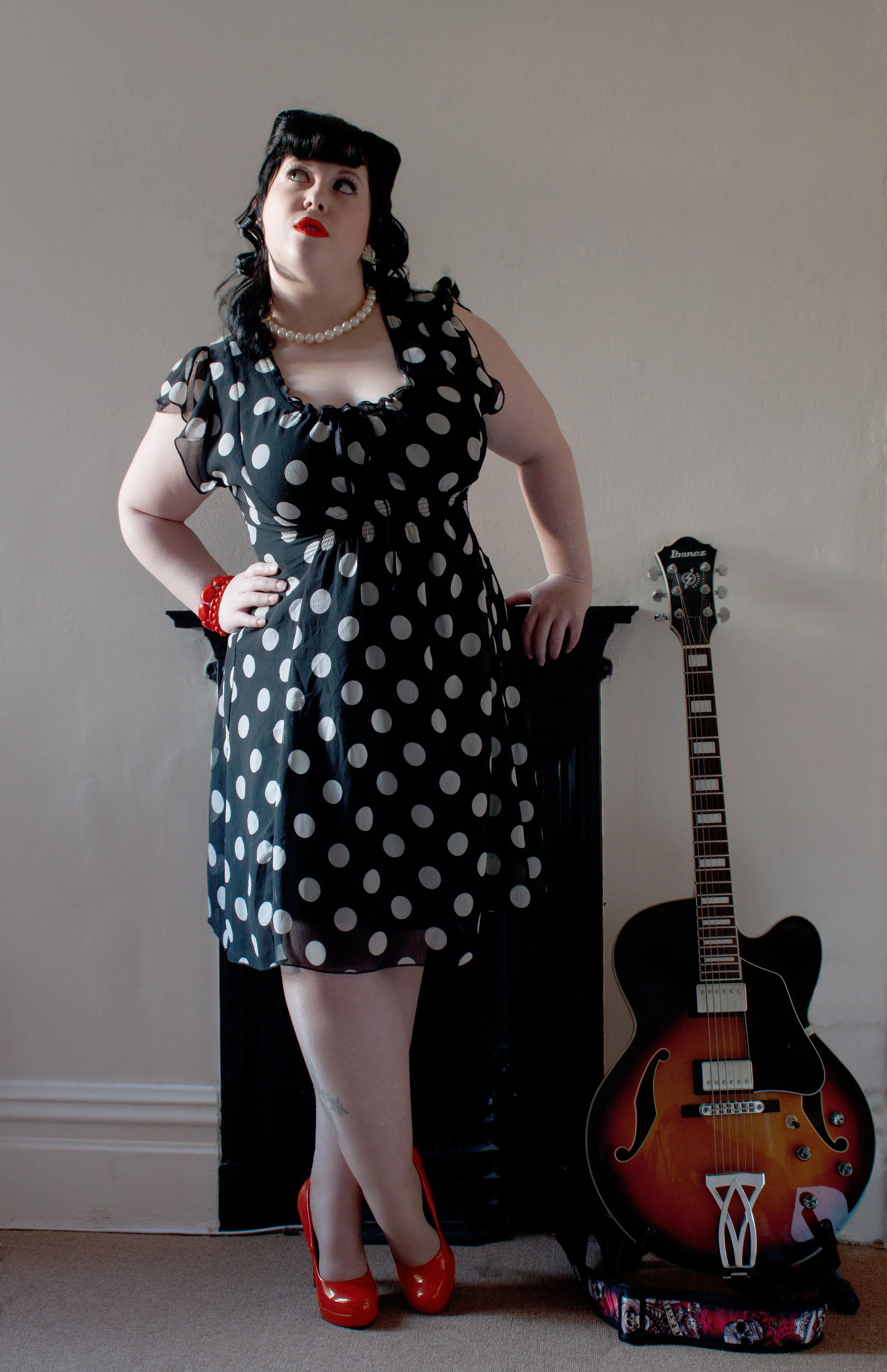 A woman with black hair, wearing a black dress with white polka dots, red shoes, a pearl necklace, and a red bracelet, stands near a black fireplace mantel with her hand on her hip. There is a sunburst electric guitar leaning against the wall beside 
