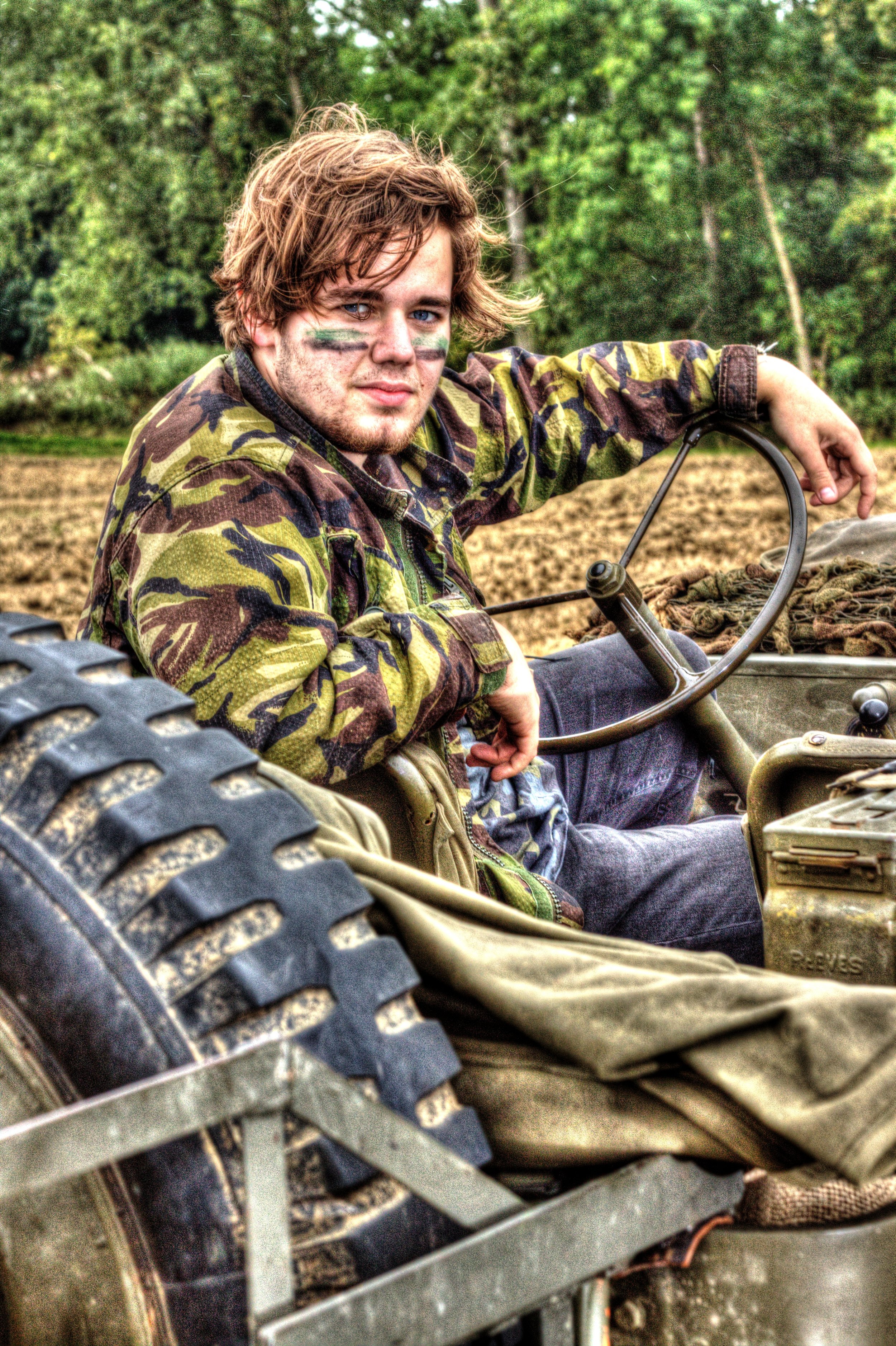 A young man with face paint and camouflage clothing sitting on a military vehicle in a rural outdoor setting with trees in the background.