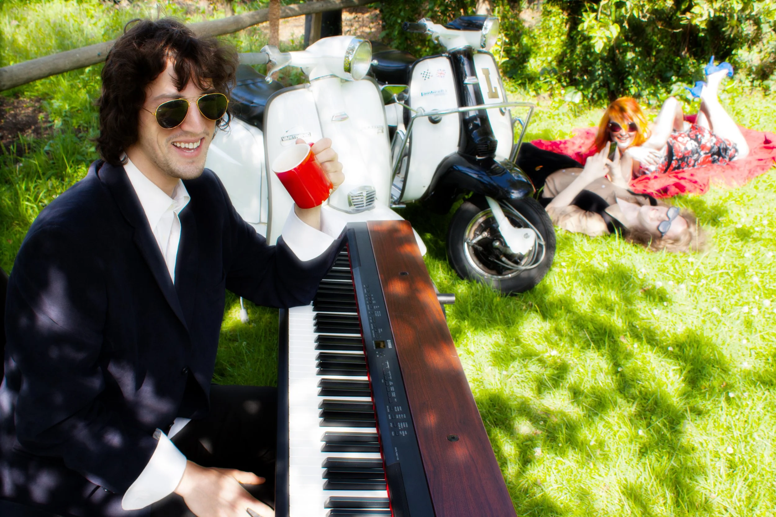 A man in a black suit and sunglasses sitting at a keyboard outdoors, holding a red mug and smiling. In the background, three women are lying on the grass, two of whom are wearing sunglasses and making peace signs, with a black scooter, two white scoo