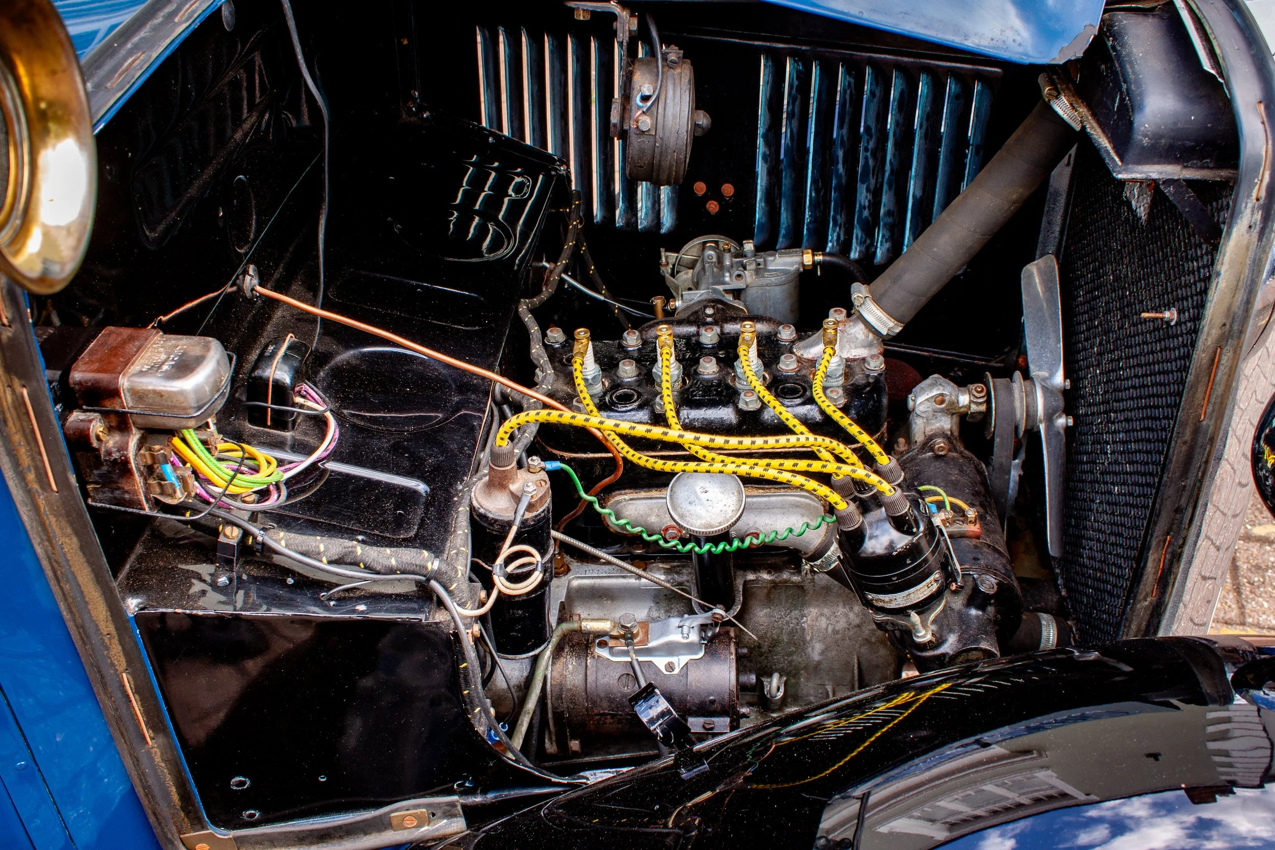 A vintage car engine bay with black components, yellow spark plug wires, and a radiator visible at the front.