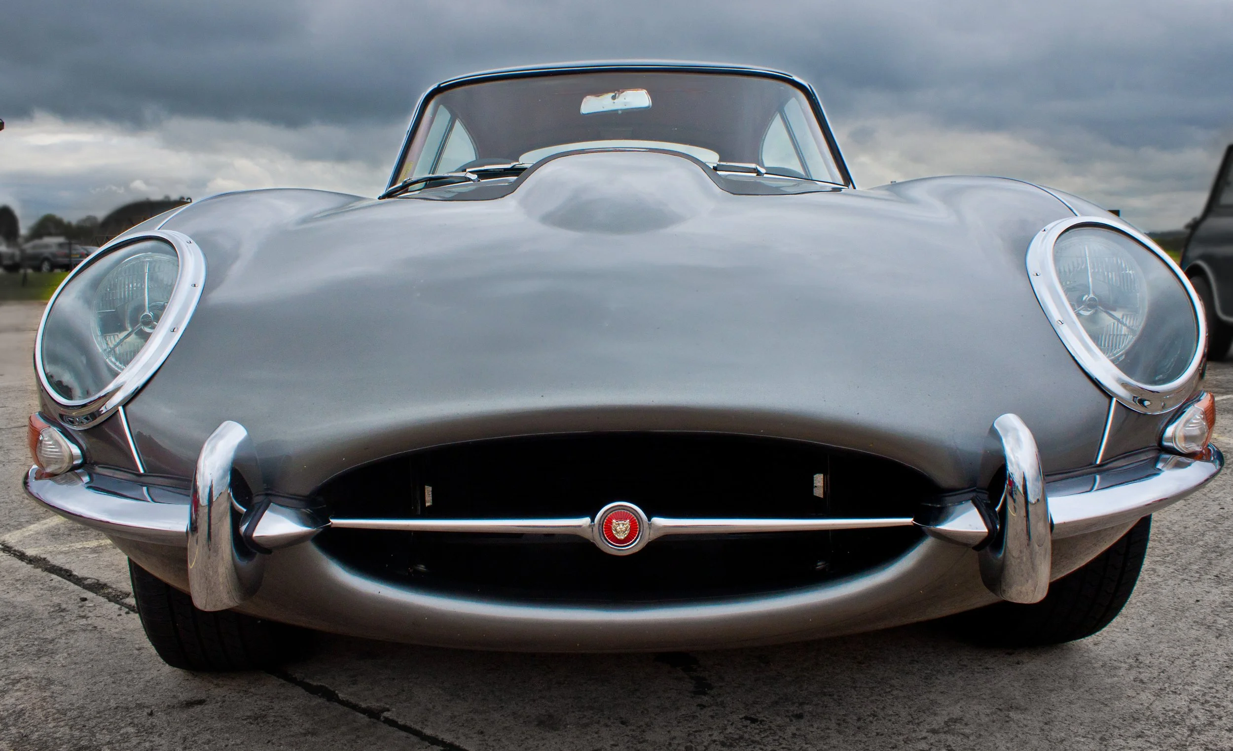 Front view of a classic silver Jaguar E-Type sports car with distinctive round headlights, chrome bumpers, and a sleek aerodynamic design, parked outdoors on a cloudy day.