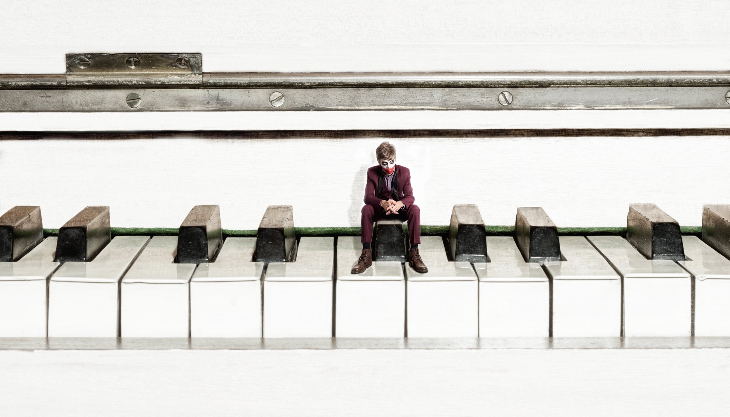Miniature person dressed as the Joker sitting on piano keys in front of a white background.