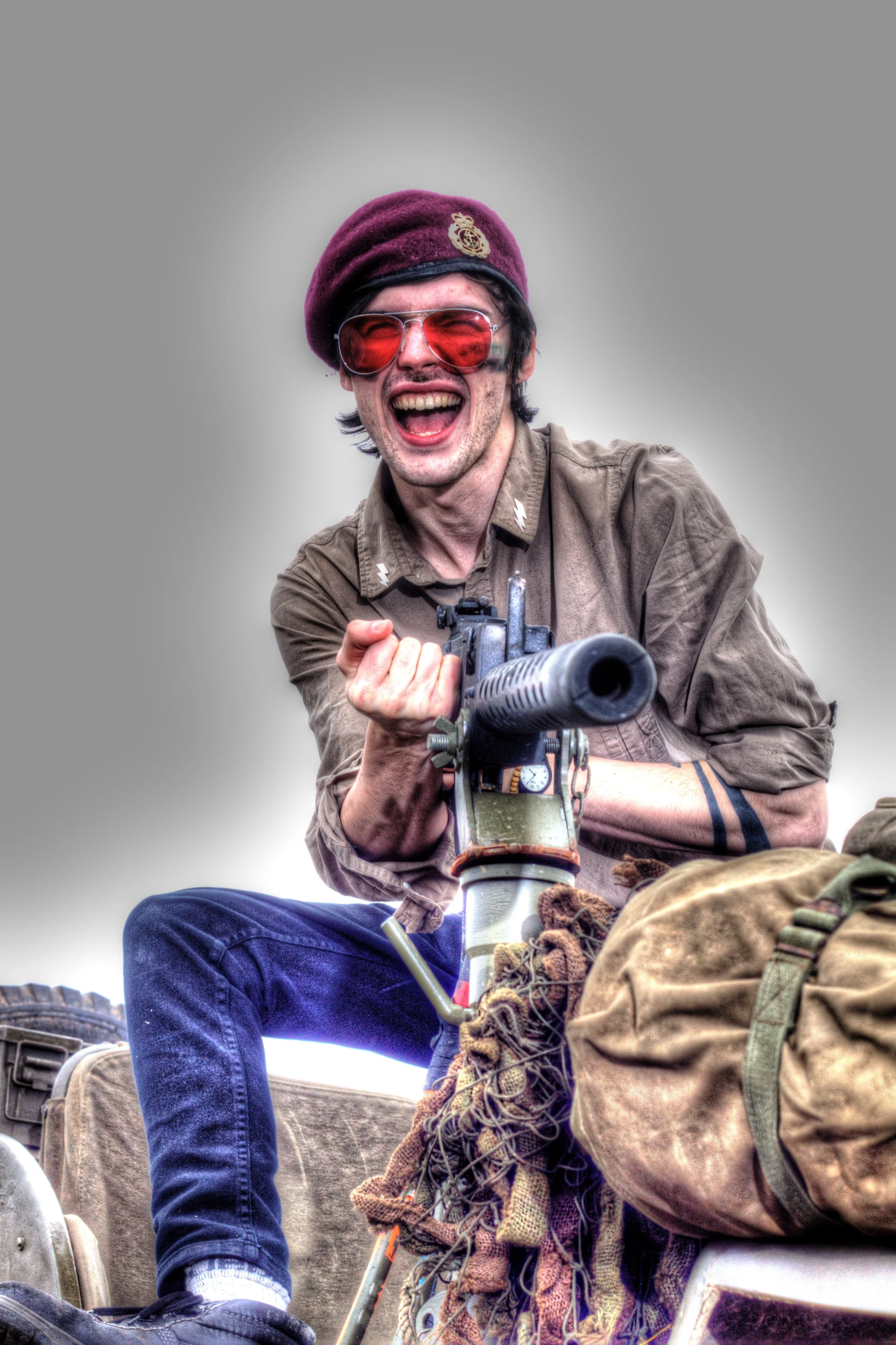 A man wearing sunglasses, a maroon beret, and a military-style shirt, smiling and pointing a gun forward while sitting on a military vehicle.