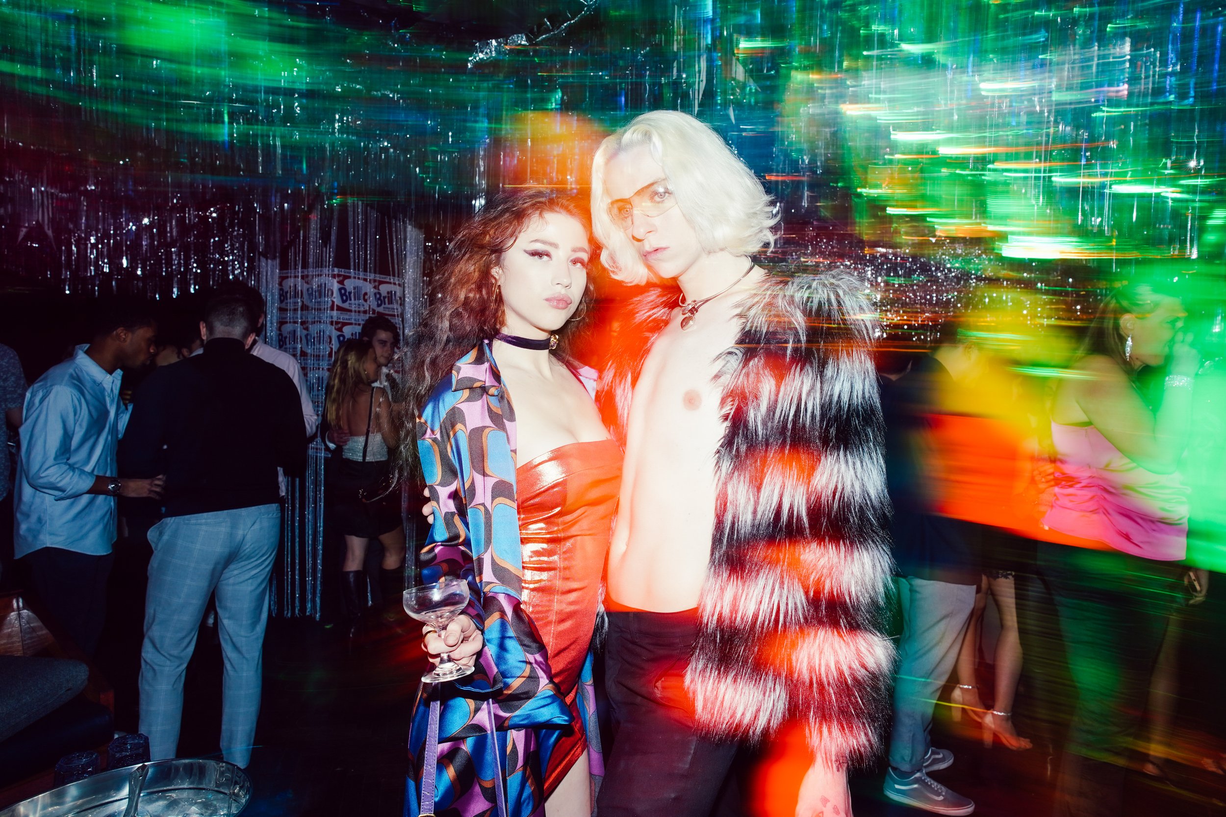 Two fashionable people posing at a lively party with colorful lights and other guests in the background.