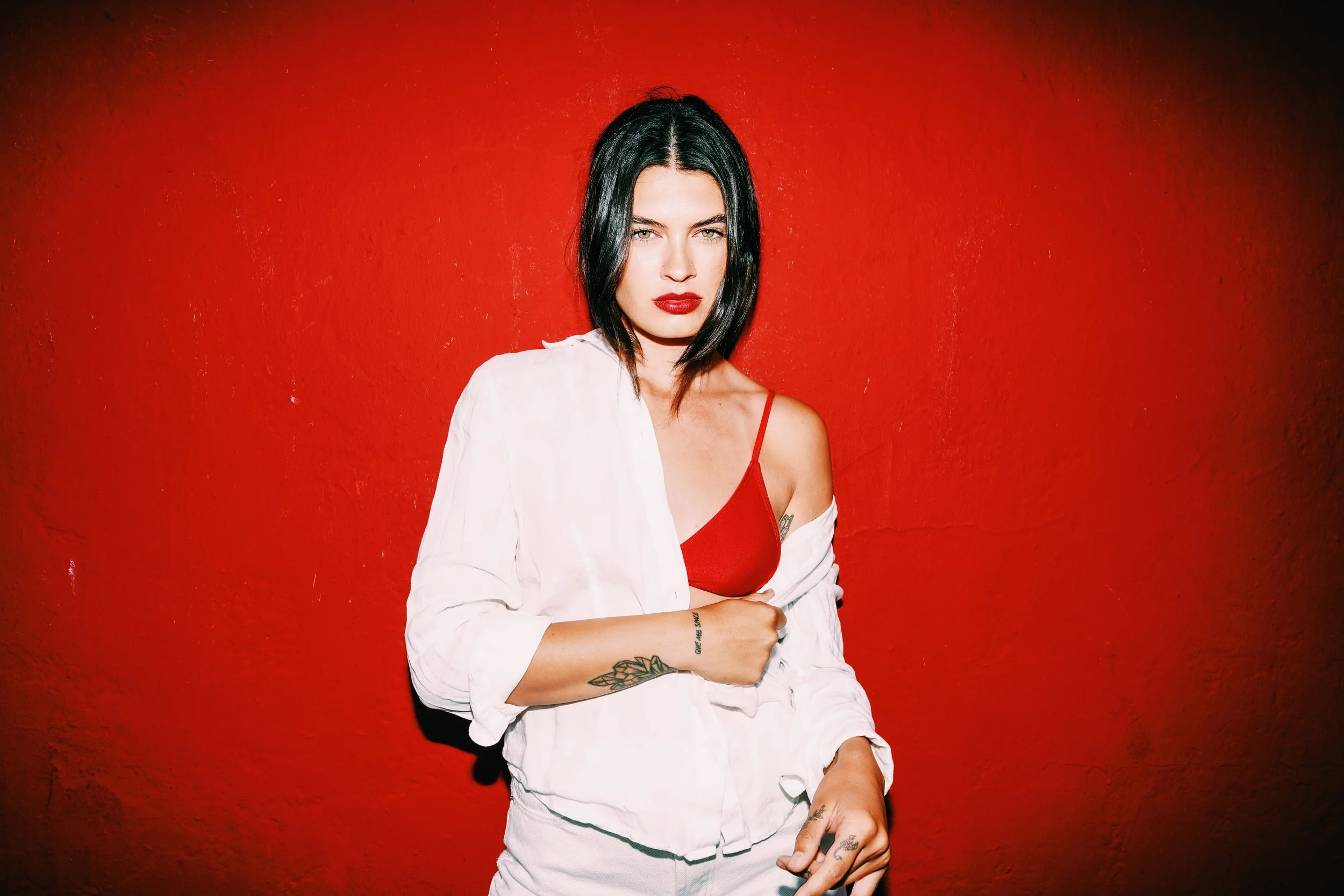 A woman with black hair and red lipstick standing against a red wall, wearing a red bra and a white shirt draped off her shoulders, with tattoos visible on her arm and hand.