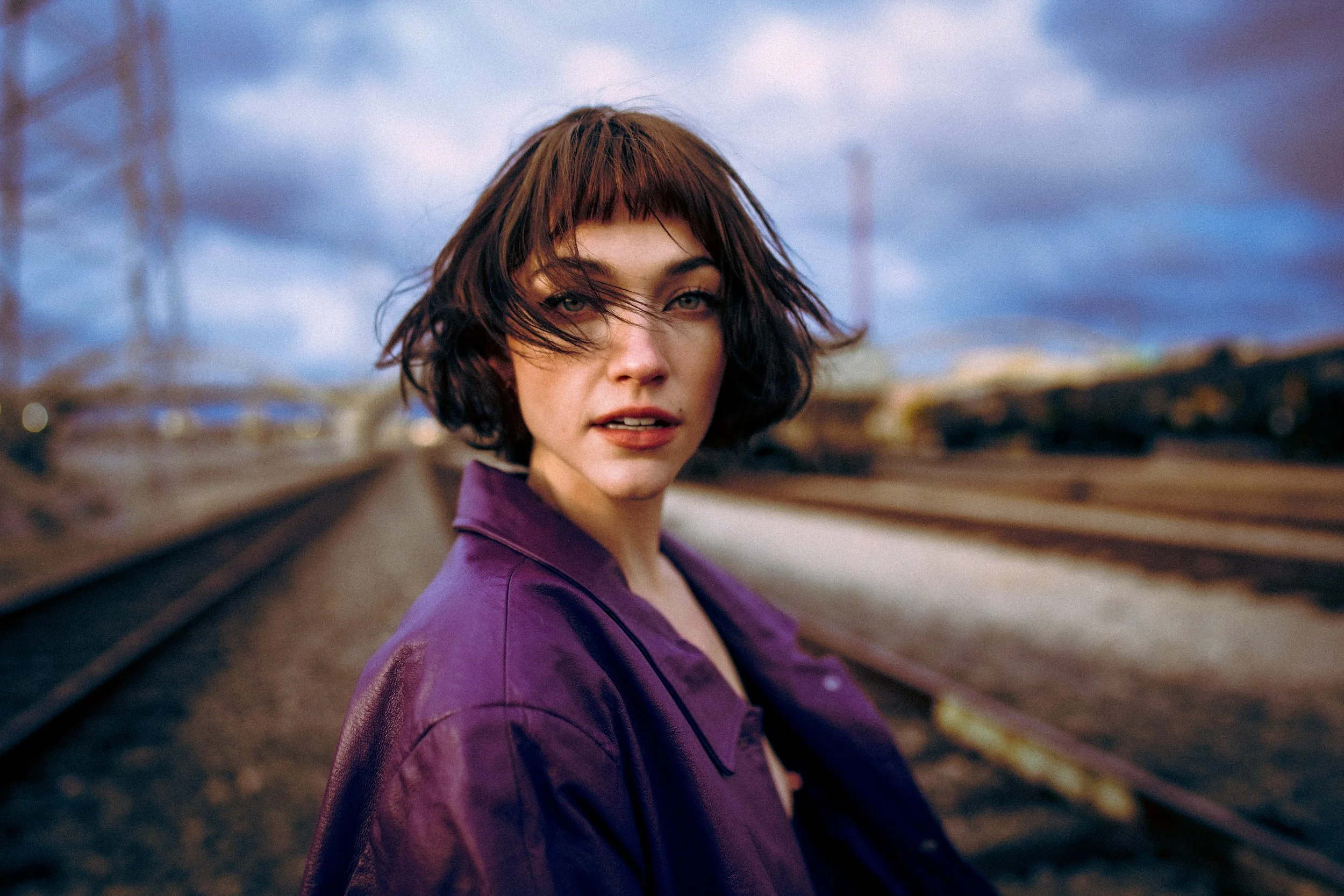 A woman with short brown hair and bangs standing on railroad tracks outdoors during overcast weather.